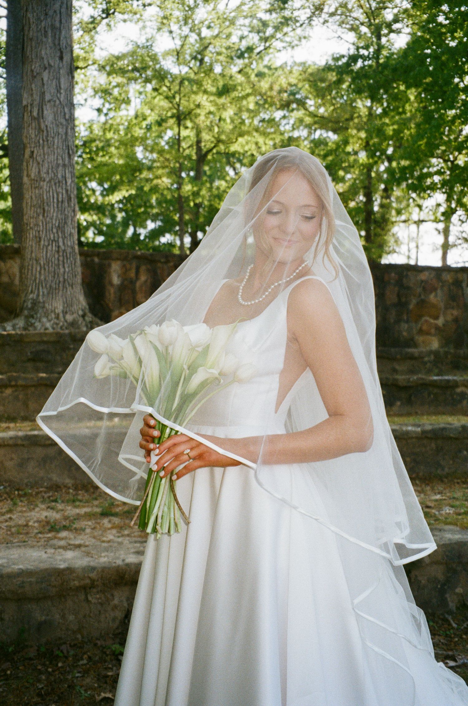 Smiling bride in white dress and veil looking down at calla lily bouquet outdoors.