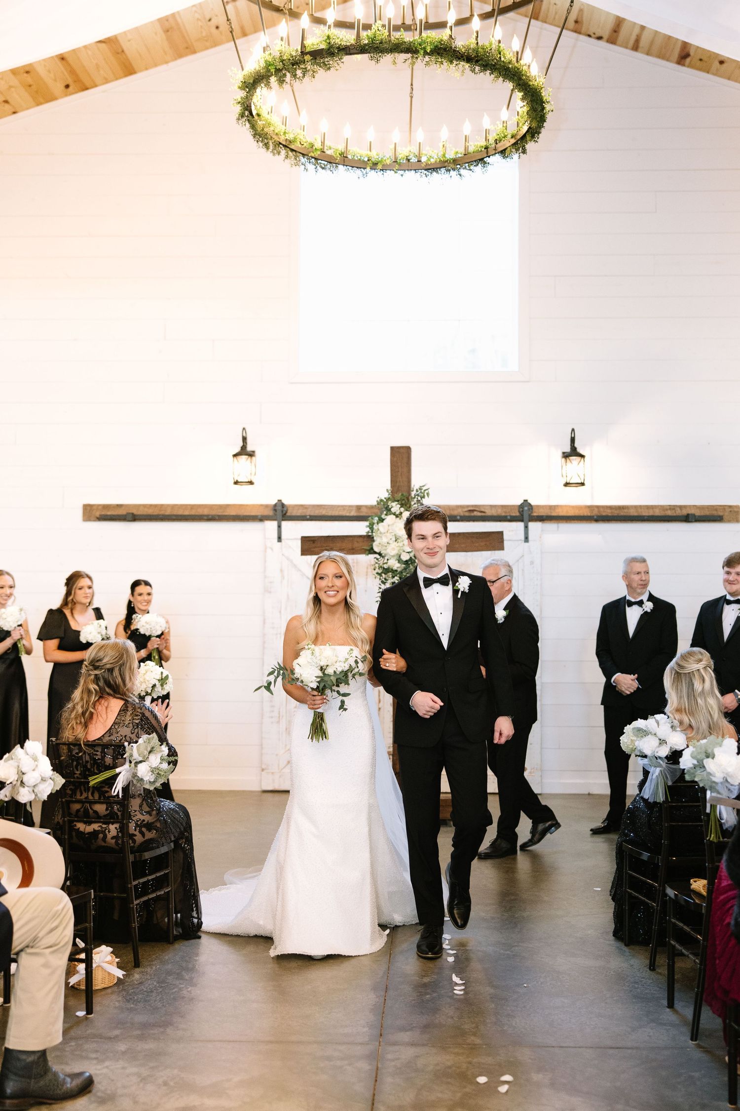 Couple walks down aisle under rustic chandelier in modern white barn venue.
