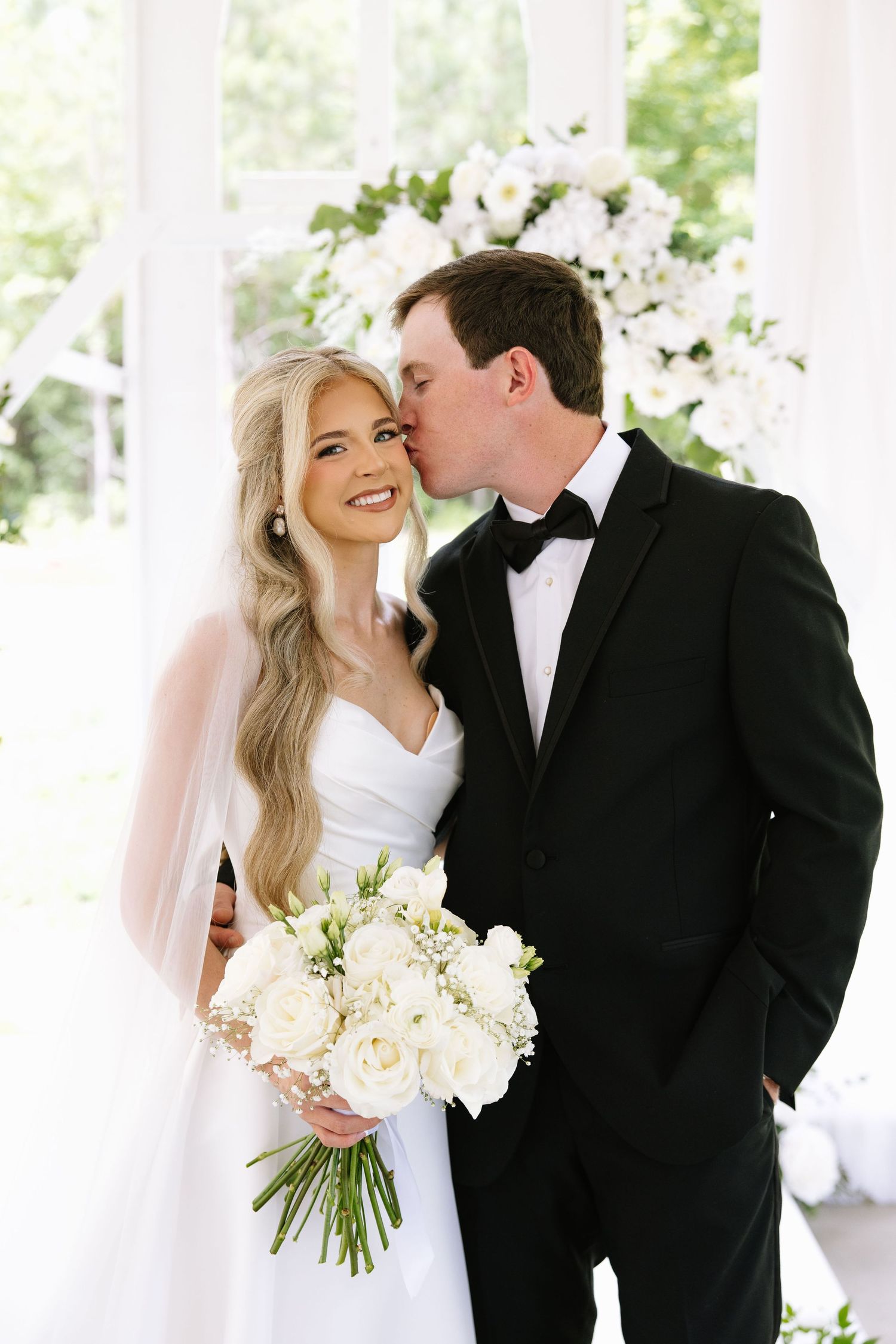 Groom in black tuxedo kissing bride on cheek as she smiles holding white rose bouquet at elegant wedding ceremony.