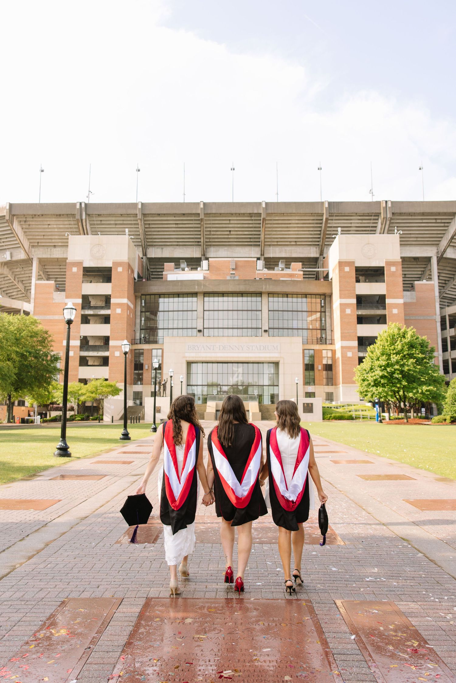Three female graduates in black gowns with red and white stoles walk toward a large university stadium.