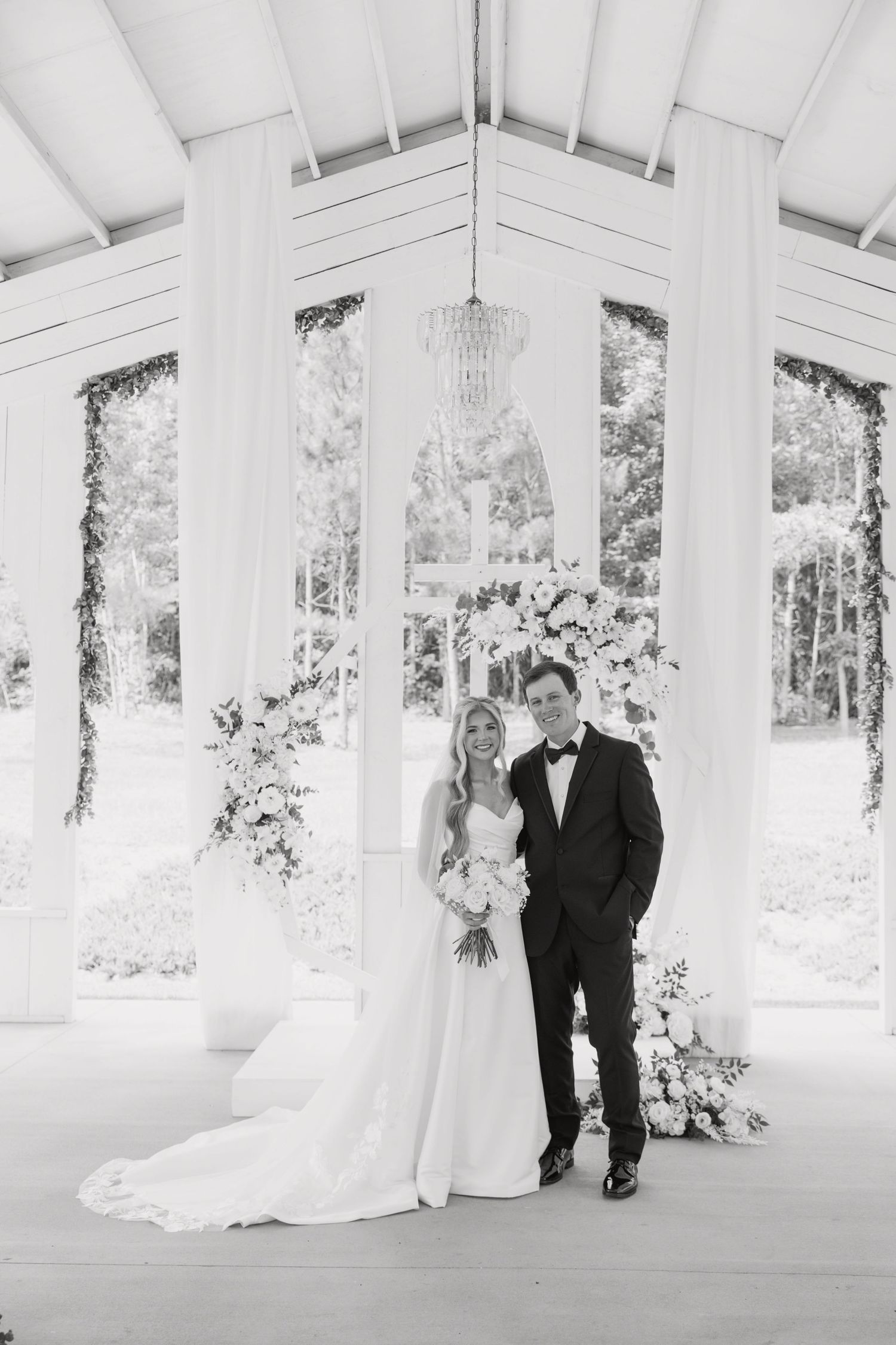 Black and white photo of a bride and groom posing under an elegant white pavilion adorned with floral arrangements.