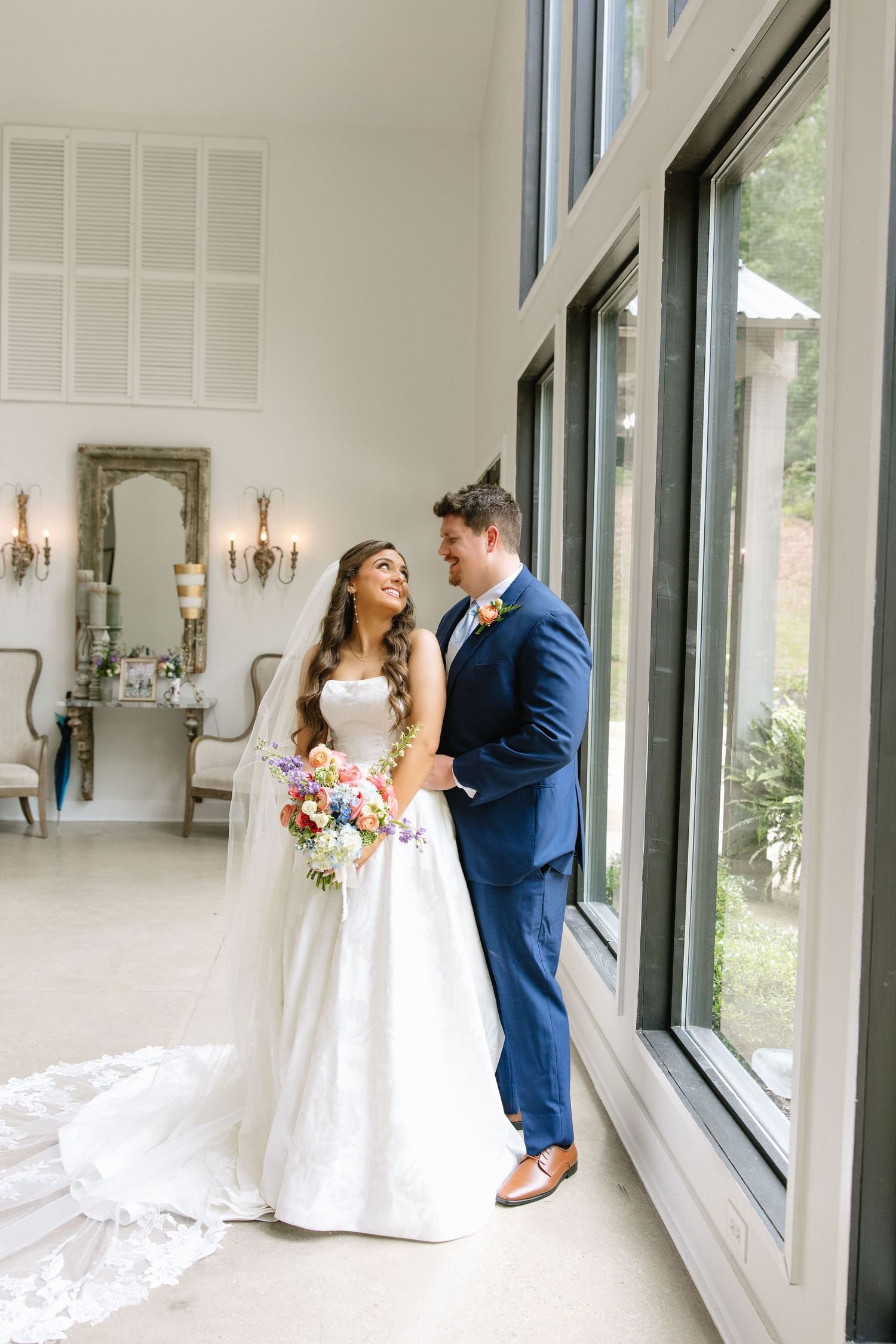 Bride and groom share a joyful moment by tall windows in an elegant venue, she holds a colorful bouquet in her white gown.