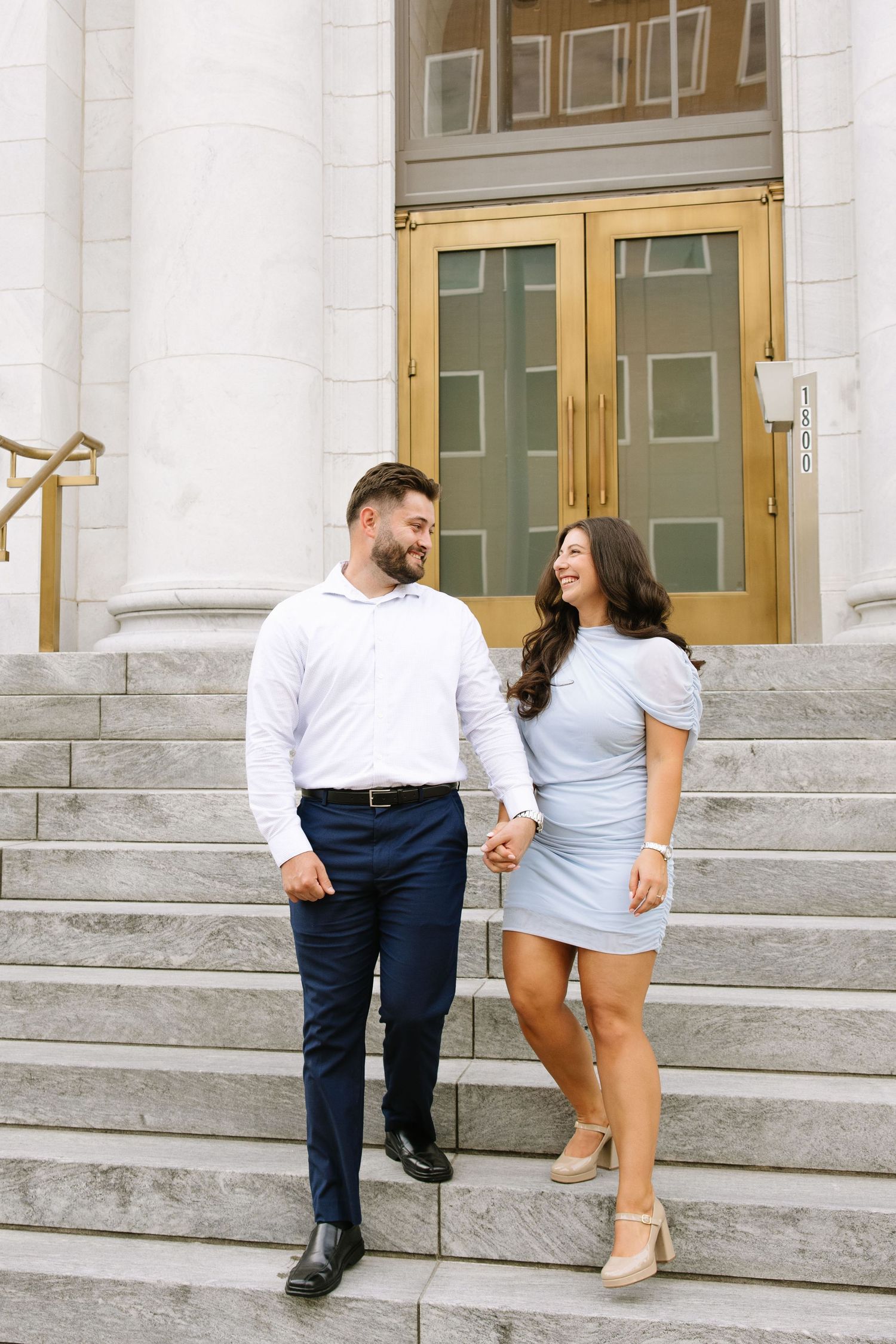 Couple holding hands and smiling at each other on grand marble steps outside a building with gold doors.