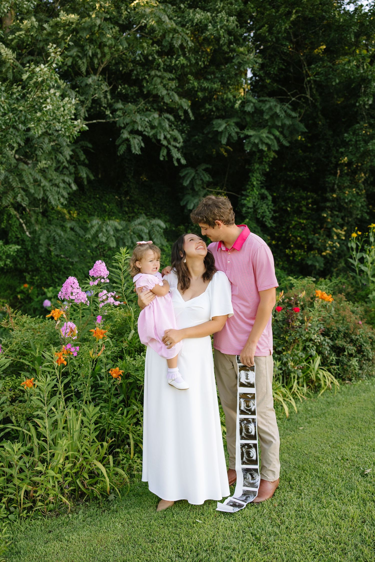 A pregnant woman in white dress holds toddler in pink while man in striped polo kisses her cheek in garden.