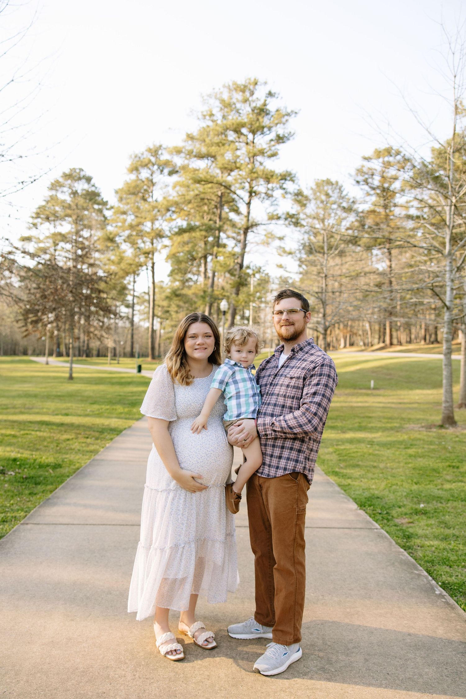 Pregnant woman in white dress with husband and toddler son posing on park path during golden hour maternity photo session.