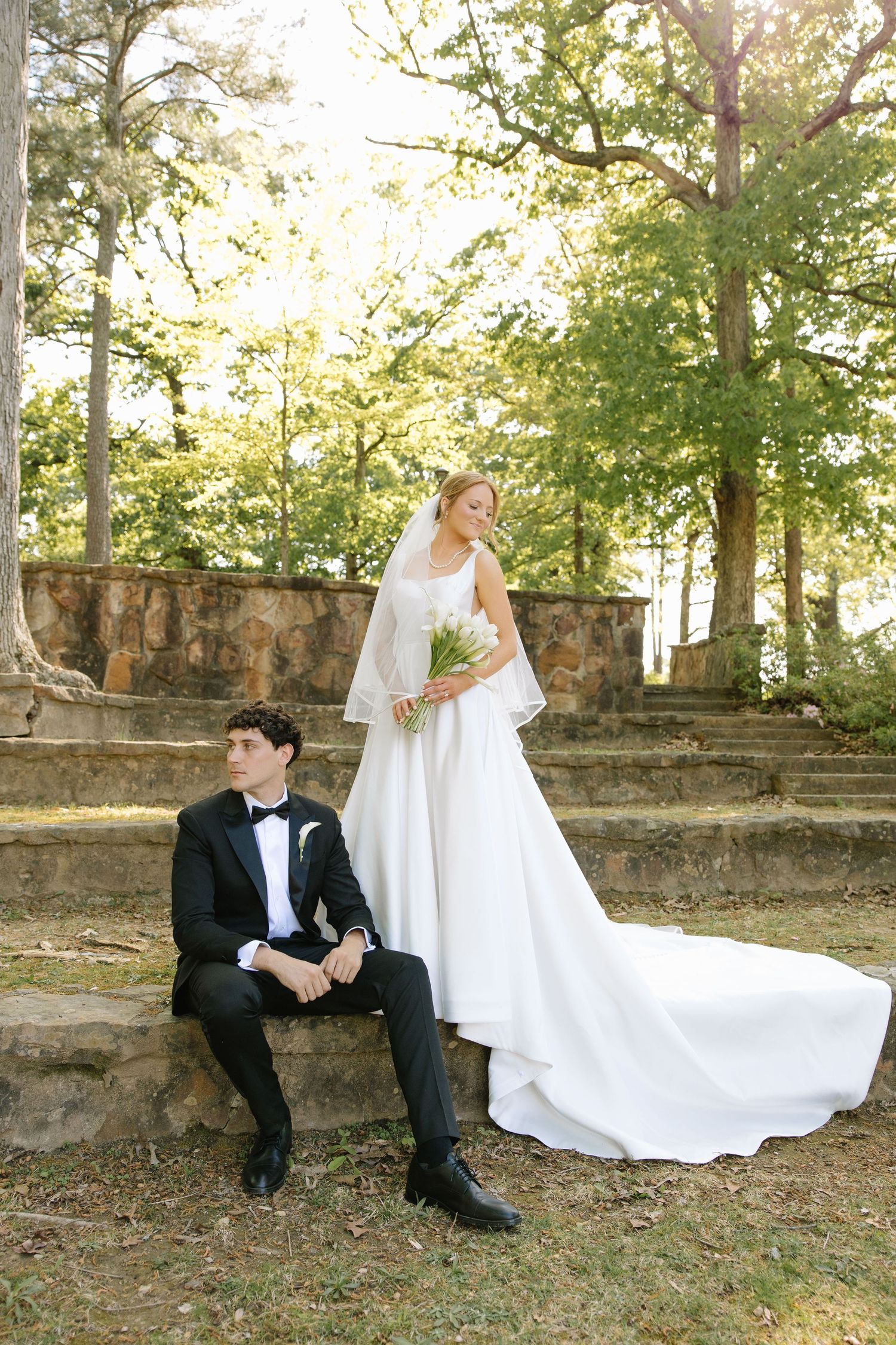 Elegant bride holding white bouquet stands beside seated groom on stone steps surrounded by lush trees.