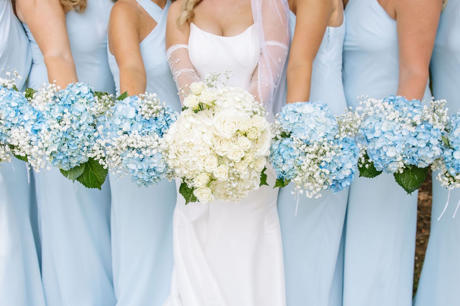 Bride in white gown holding white hydrangea bouquet surrounded by bridesmaids in light blue dresses with blue hydrangeas.