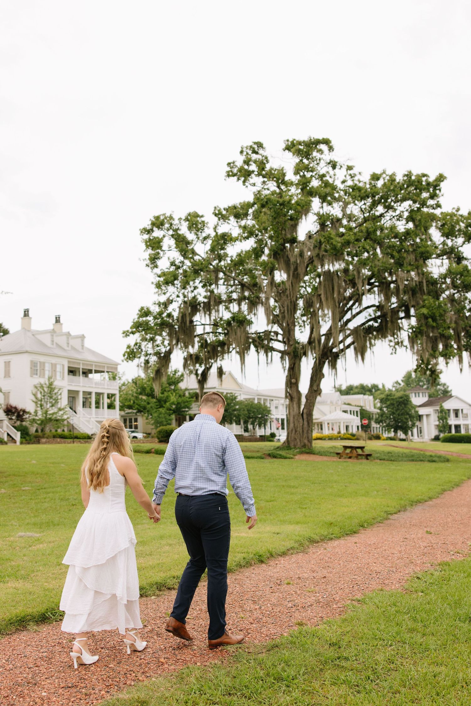 Couple holding hands walking along a path with Southern plantation homes and a moss-draped oak tree in the background.