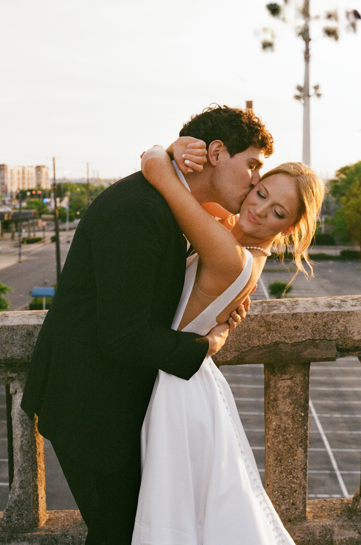 Groom in black tux embracing bride in white gown on outdoor terrace at sunset.