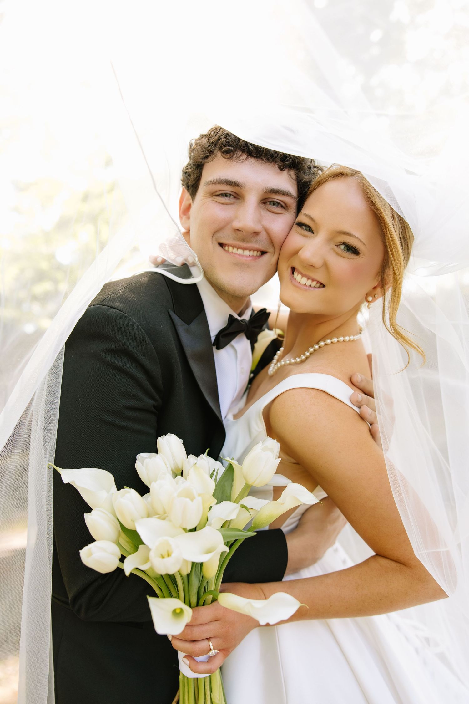 Smiling bride and groom pose under veil, she holds white calla lily bouquet wearing pearls and strapless gown.