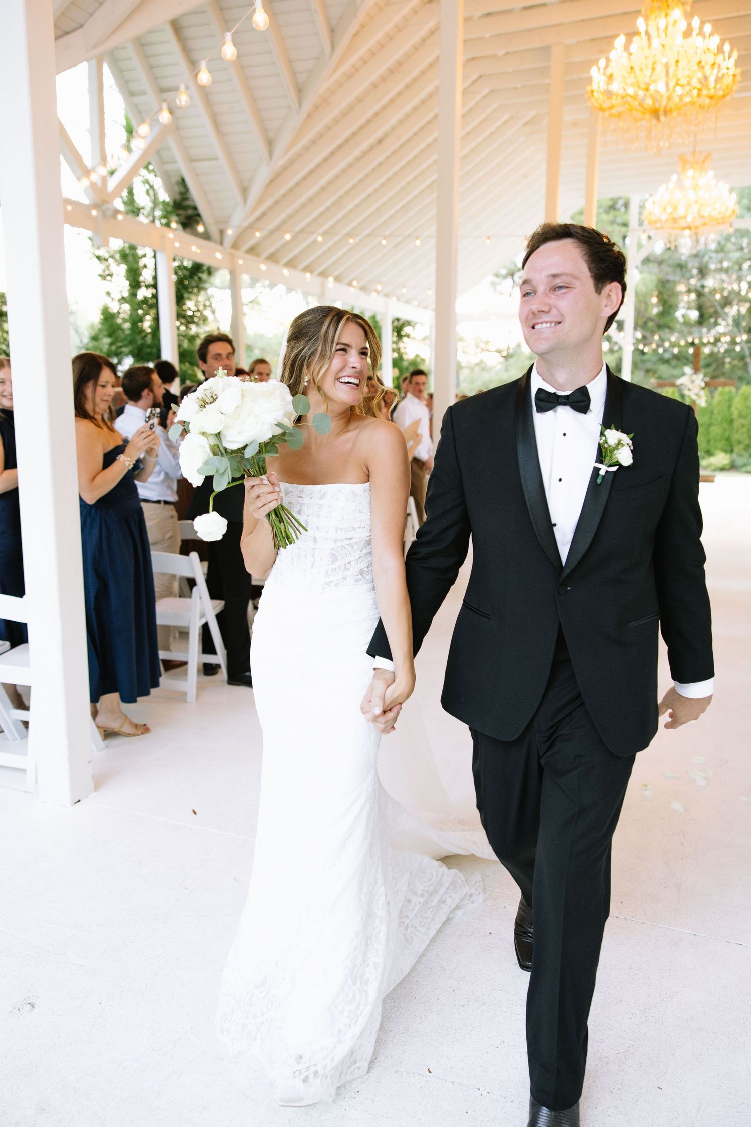 Wedding couple dances together in an elegant white reception venue with chandeliers overhead.