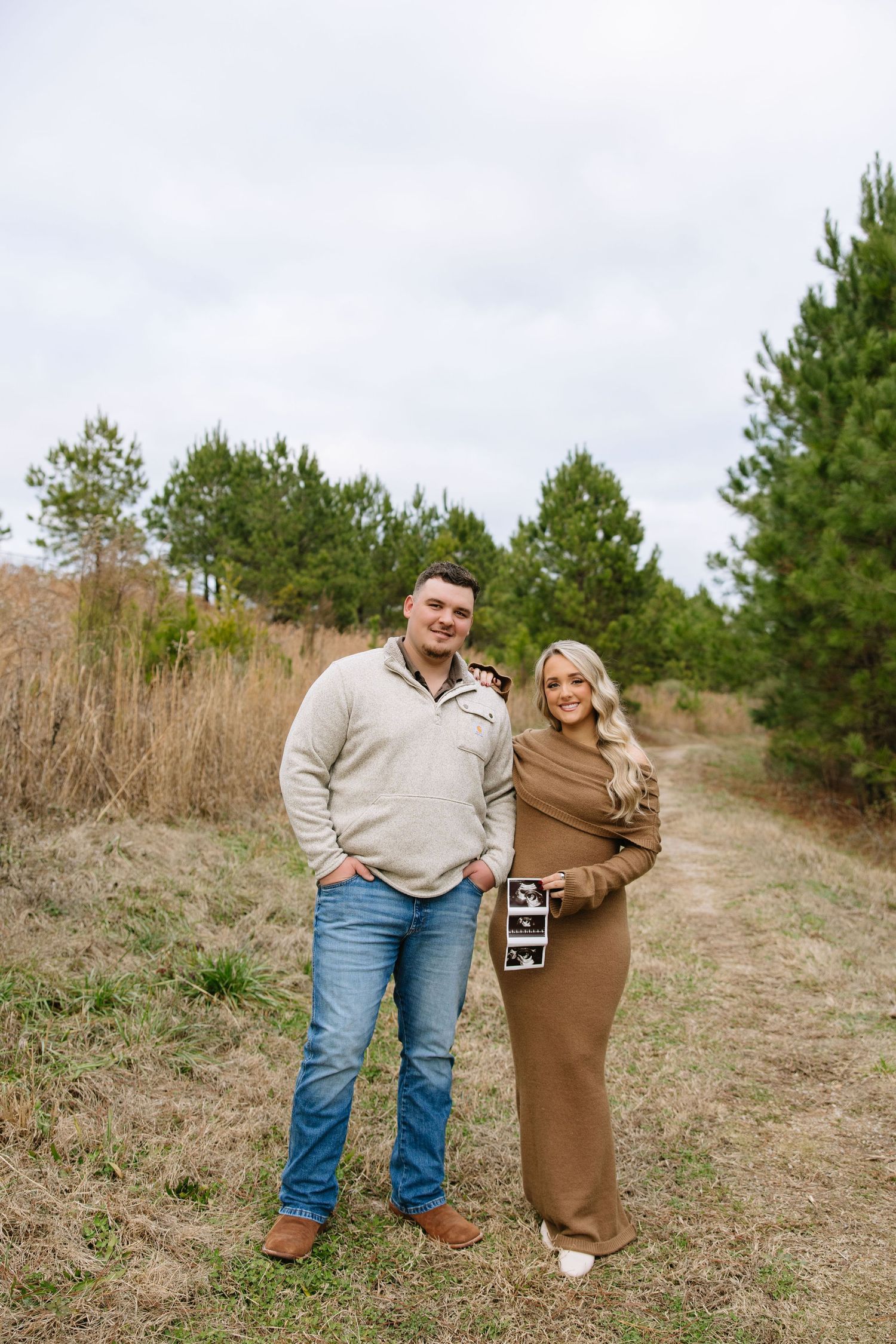 Couple smiling outdoors on a nature path, woman holding ultrasound photos, announcing pregnancy in a scenic wooded setting.