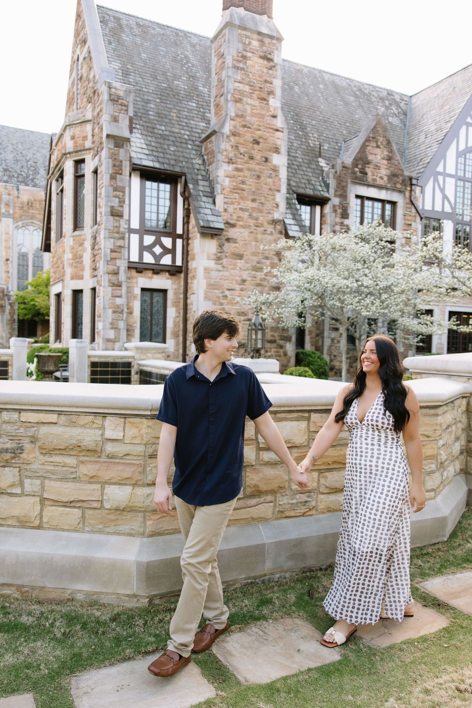 A couple holds hands and smiles while walking outside a stone Tudor-style building with blooming white trees.
