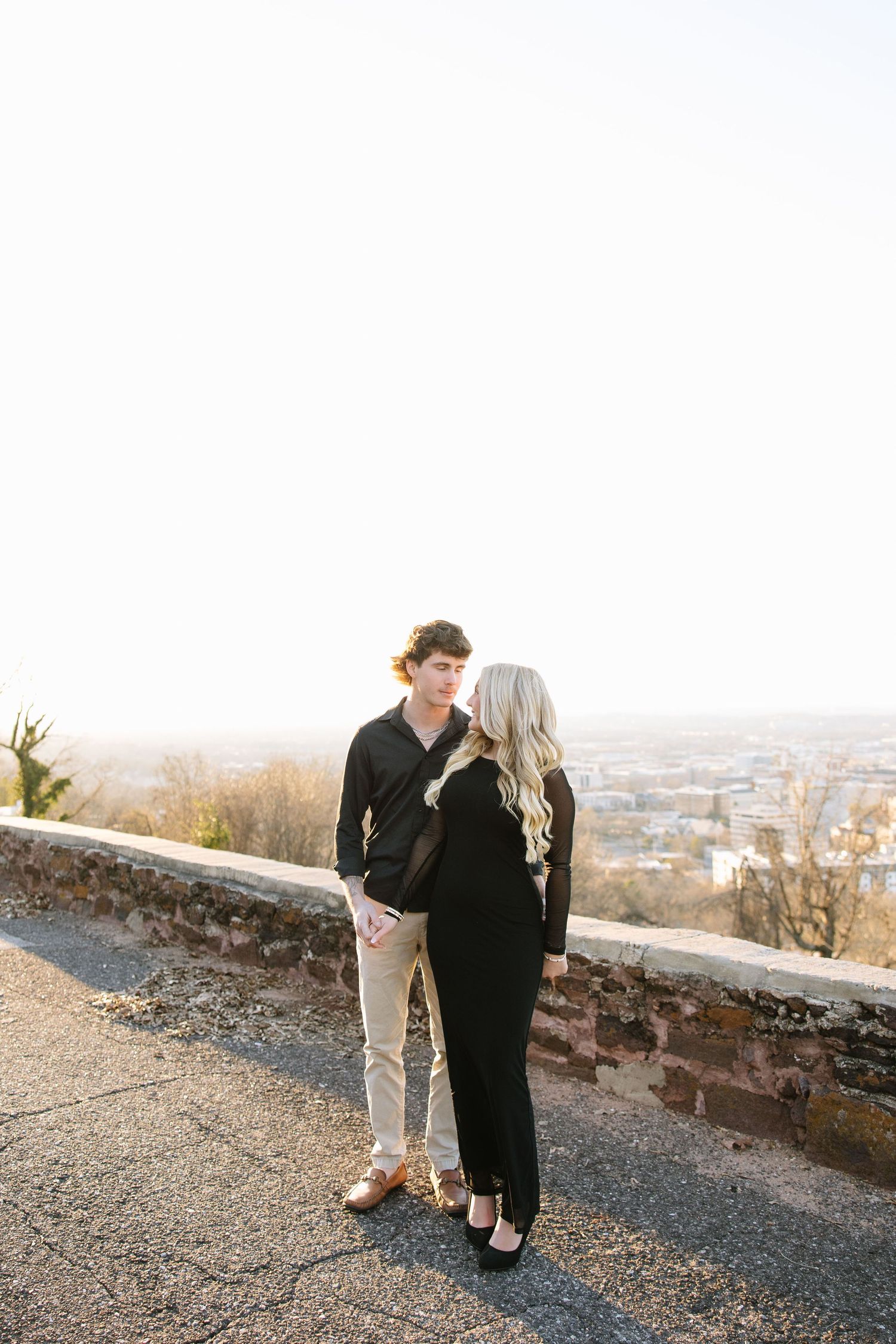 A couple in black outfits holds hands on a scenic overlook with a city view at golden hour.