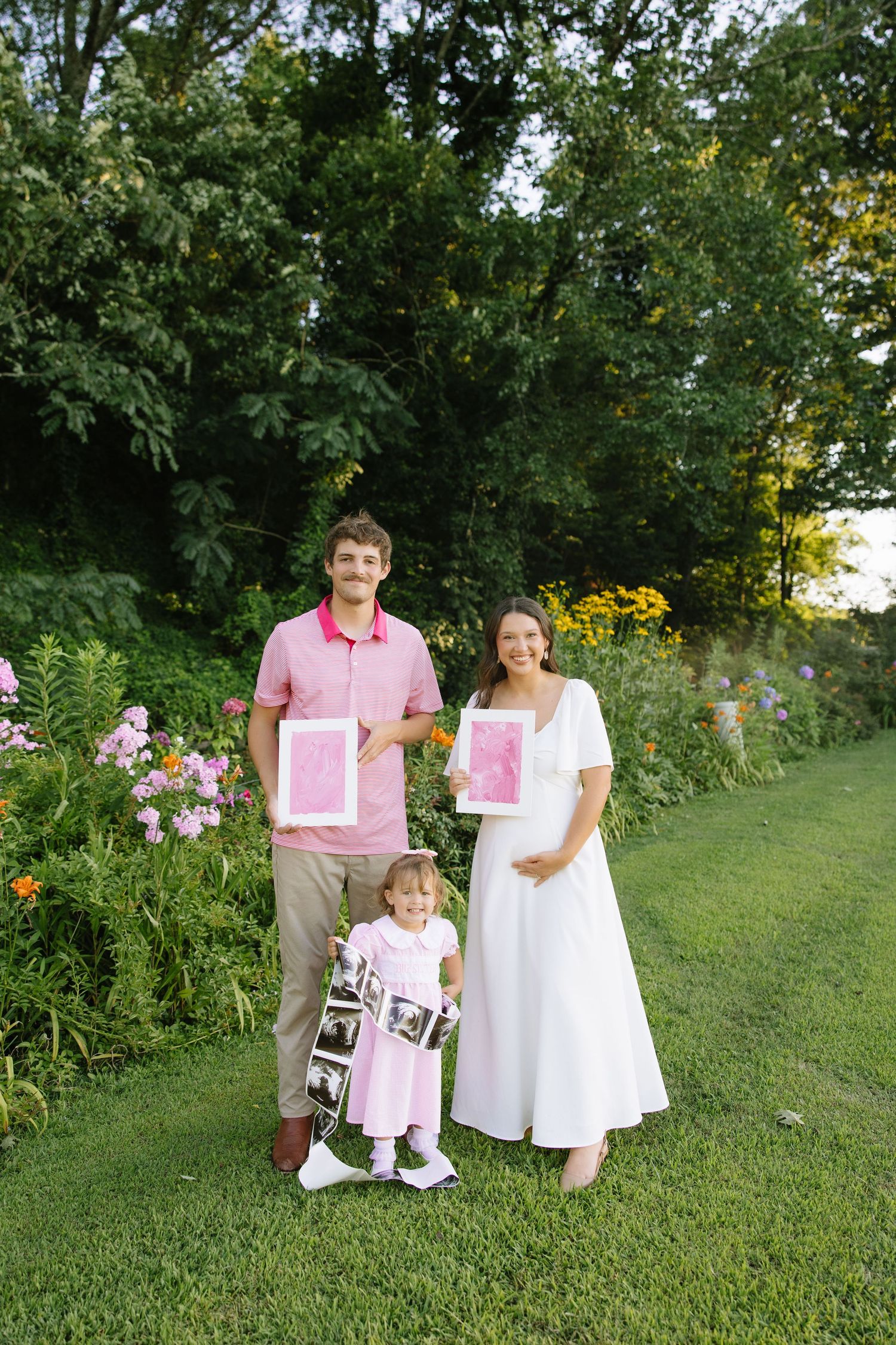 Pregnant woman in white dress and man in pink shirt hold pink gender reveal signs with toddler in garden.
