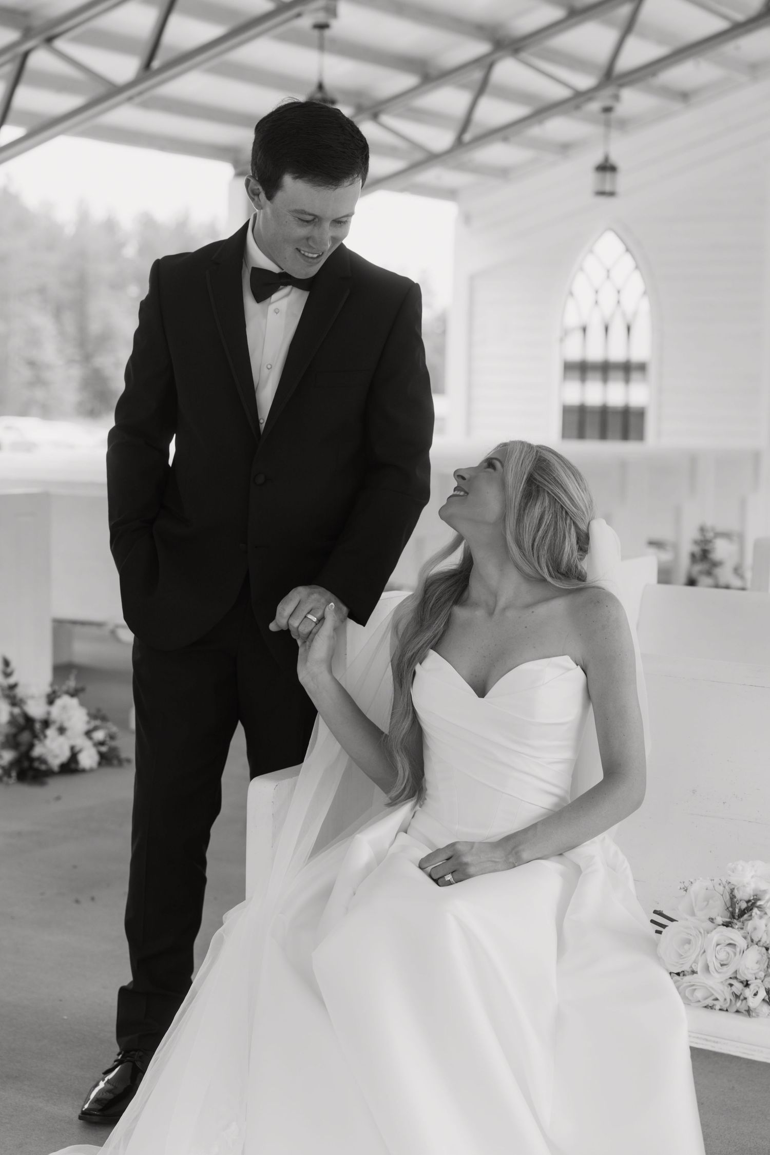 Black and white wedding photo of smiling bride in strapless gown seated, holding hands with groom in tuxedo.