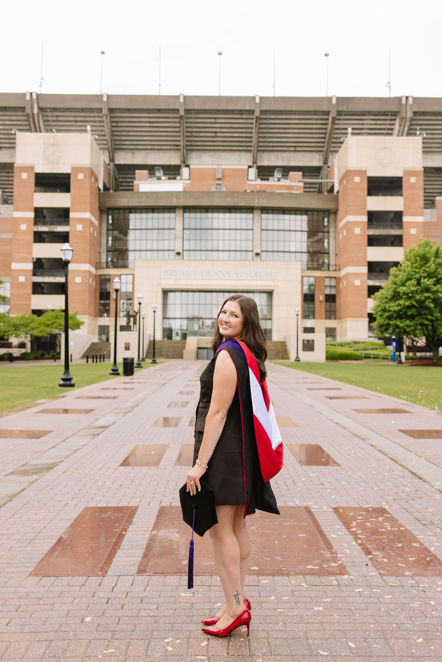 A woman in black graduation gown and red heels poses proudly in front of a large university stadium.
