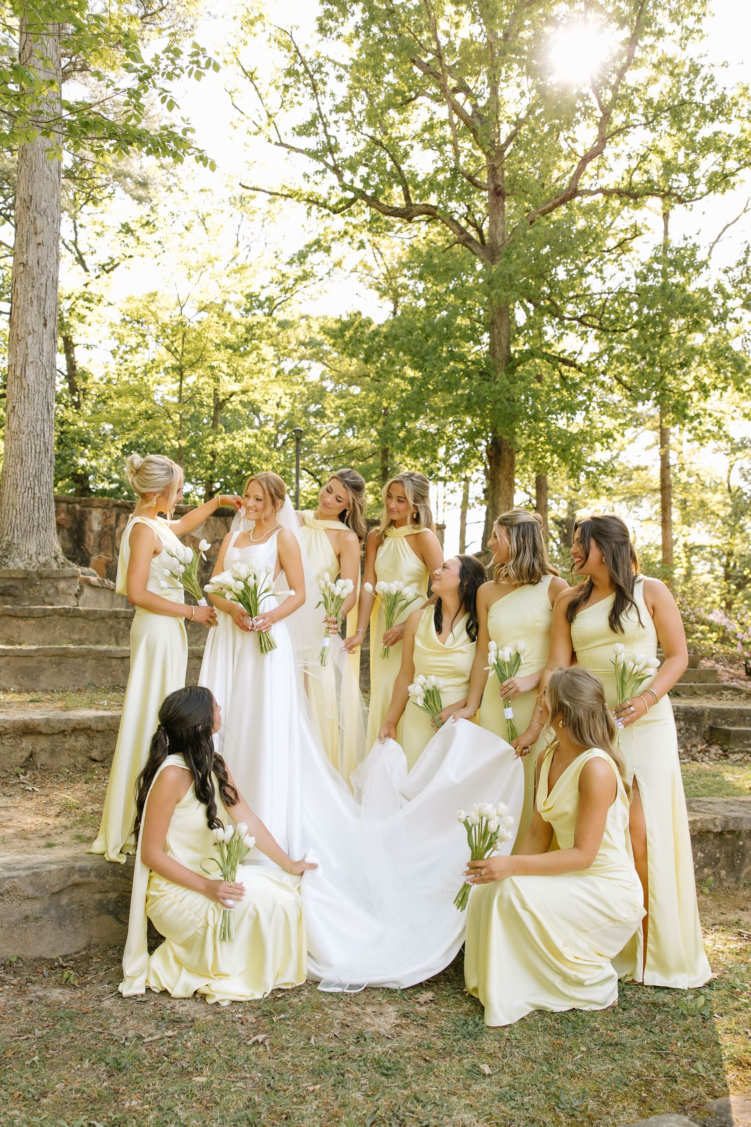 Bride surrounded by bridesmaids in yellow dresses holding white bouquets on stone steps outdoors.