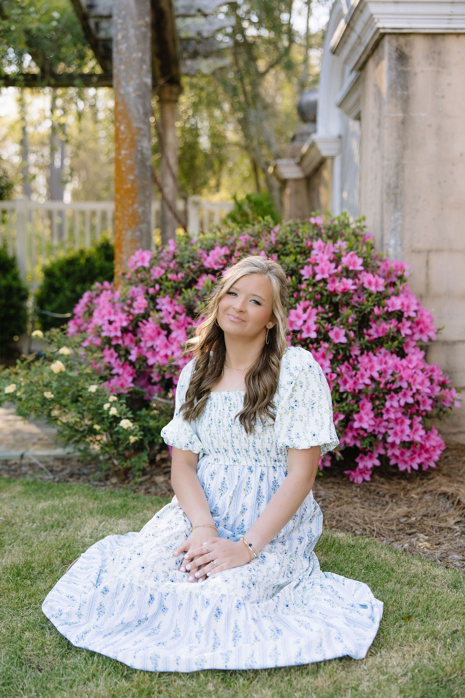 Young woman in blue floral dress sitting on grass before vibrant pink azalea bushes in a garden setting.