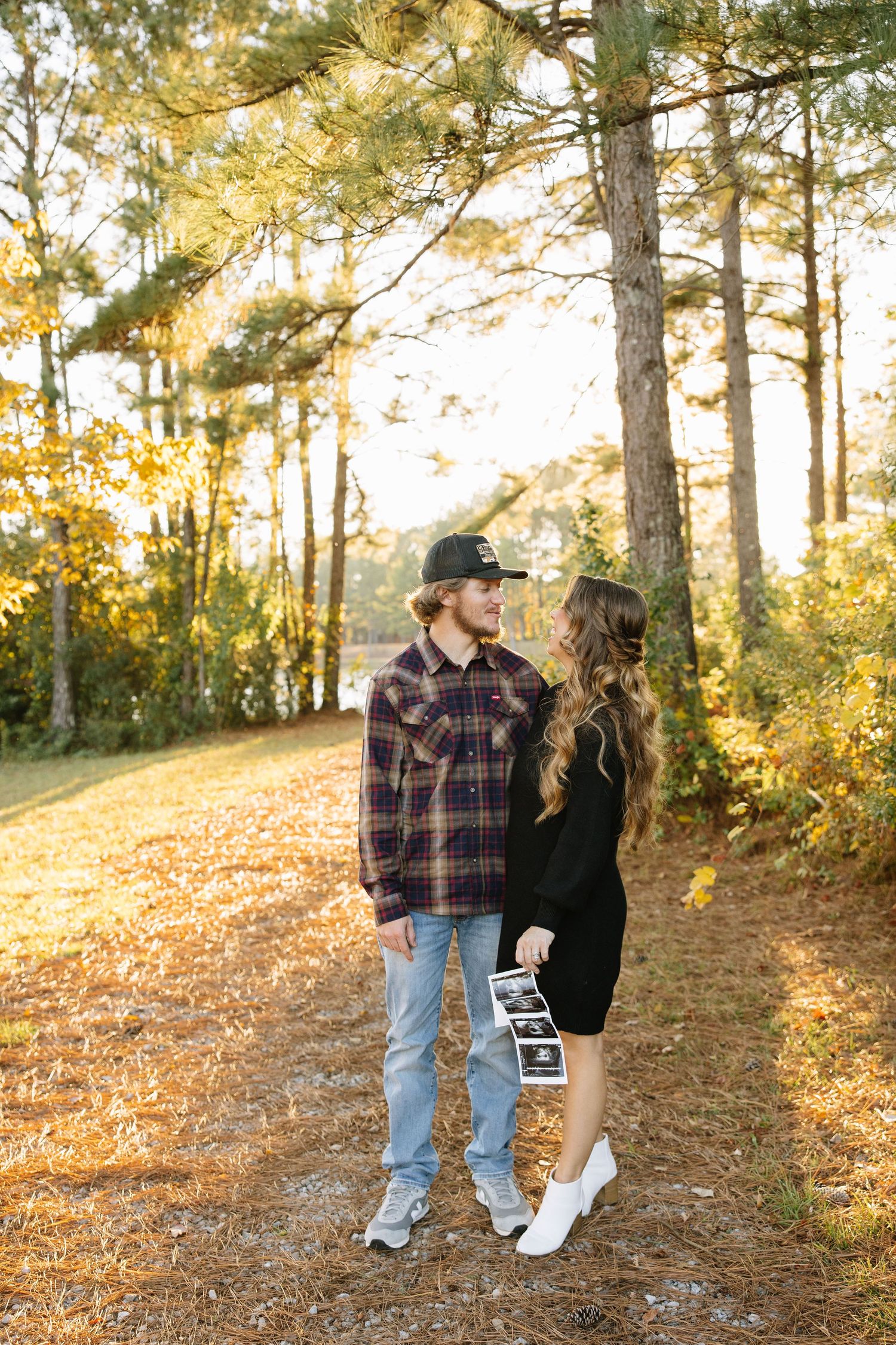 Couple holding ultrasound photos in autumn forest, pregnancy announcement in golden fall foliage.