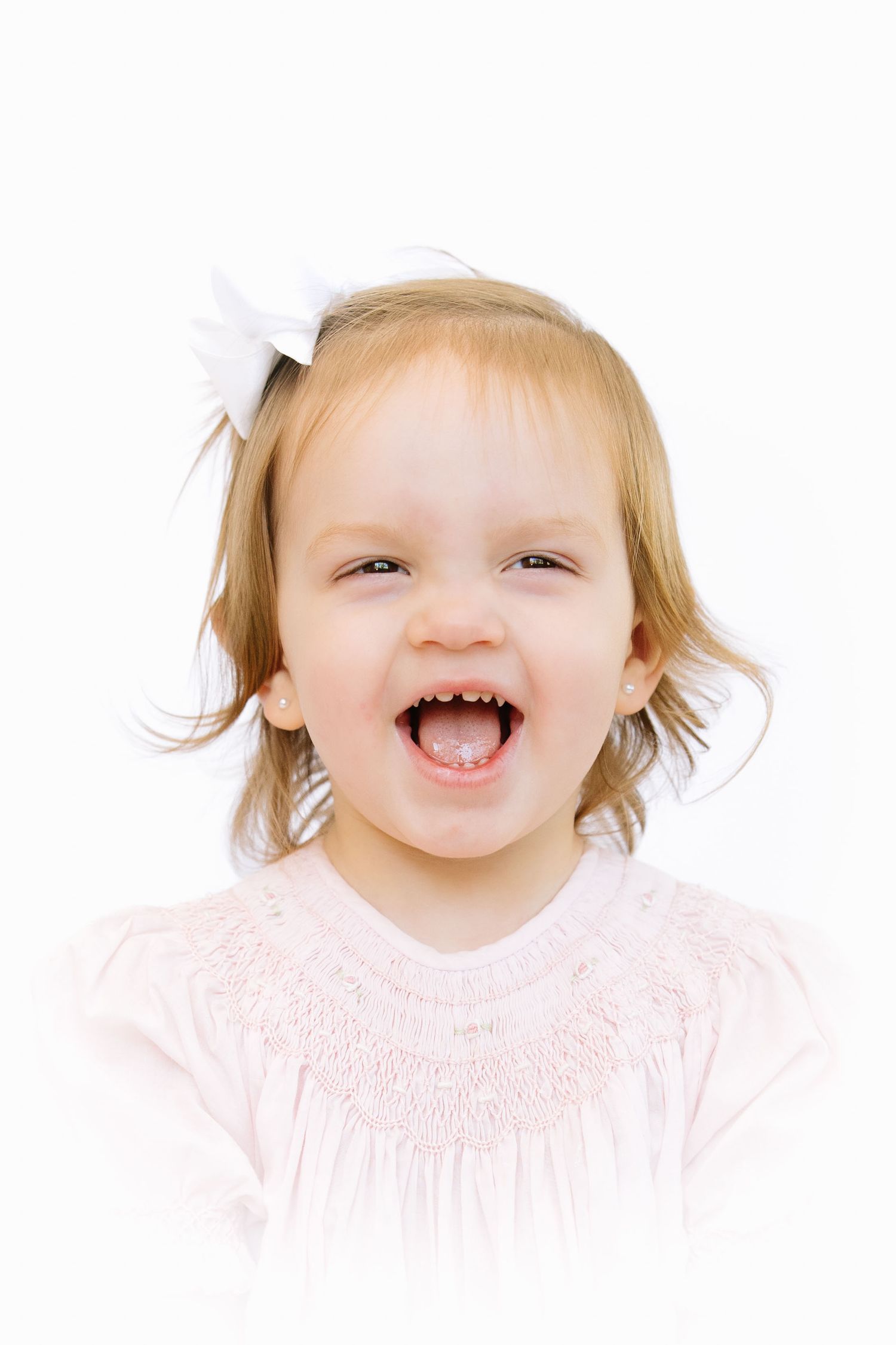 Cheerful toddler girl with white bow laughing, wearing pink smocked dress against white background.