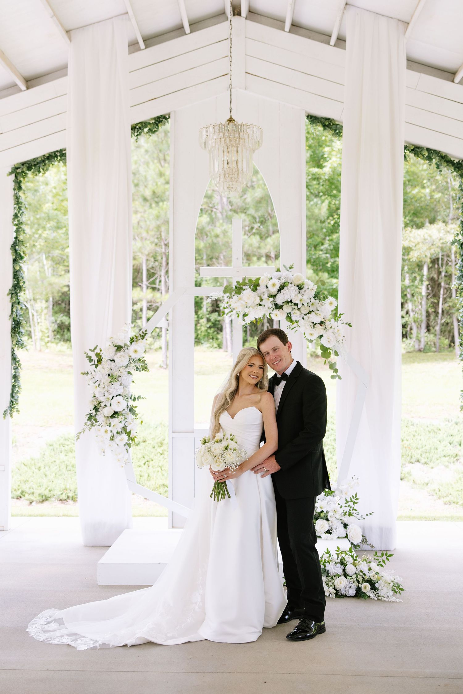 Bride and groom pose under white floral arch in elegant open-air chapel with chandelier and lush greenery backdrop.