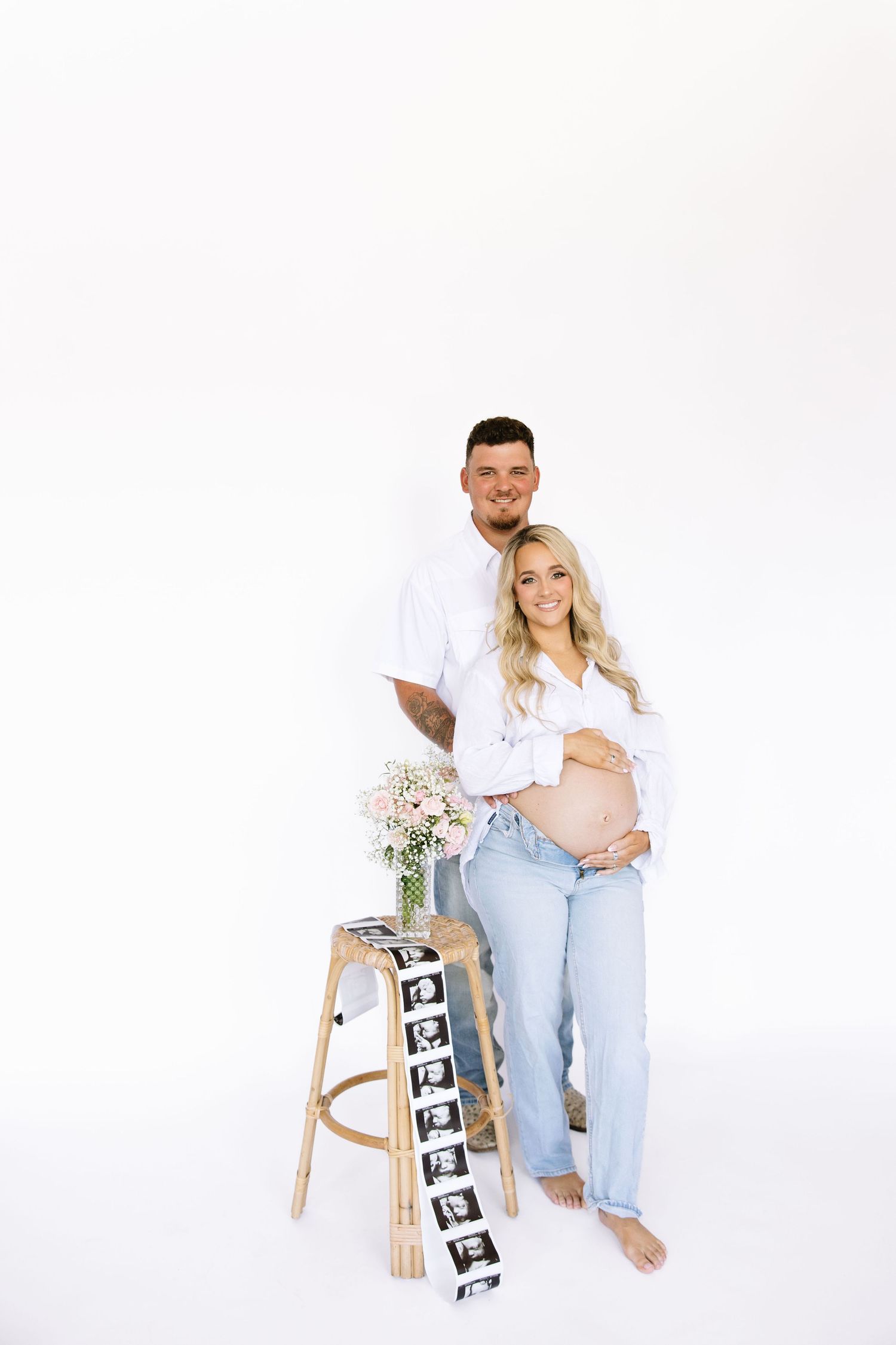 Pregnant couple in white outfits posing with ultrasound photos and flowers on a stool against white background.