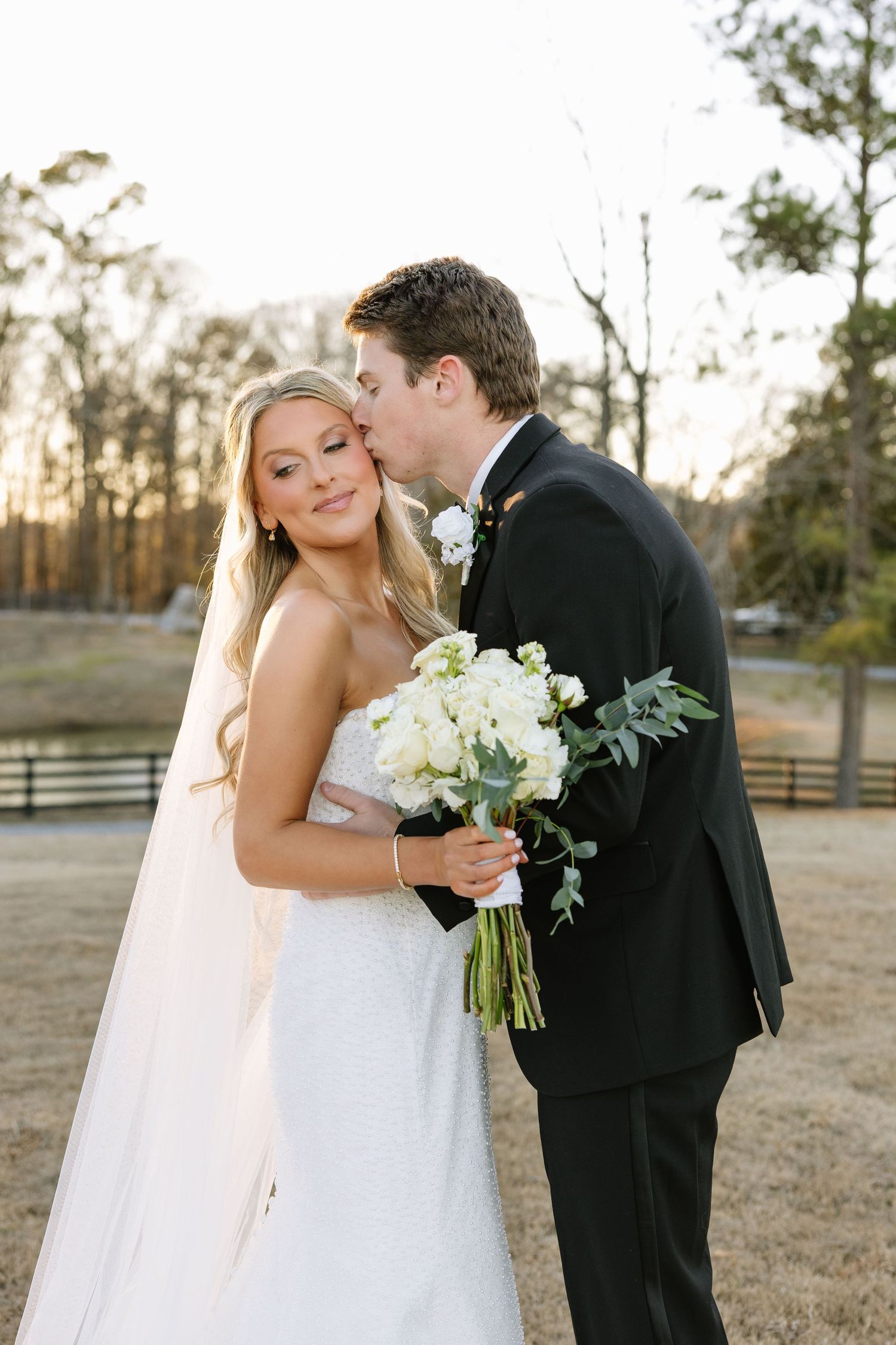 Romantic outdoor portrait of newlyweds with white bouquet at sunset.