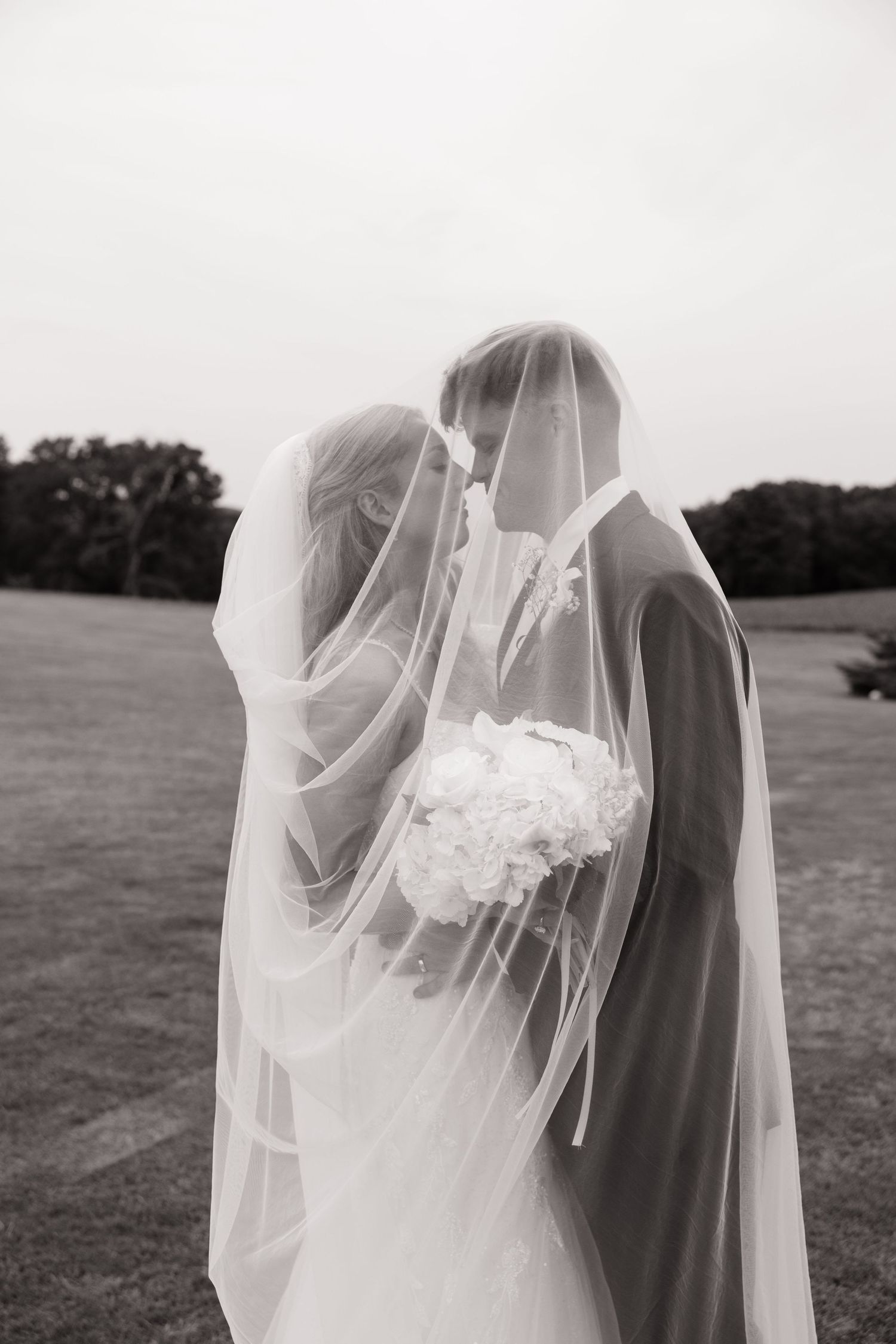 A romantic wedding couple embraces under a flowing white veil in a serene outdoor setting.