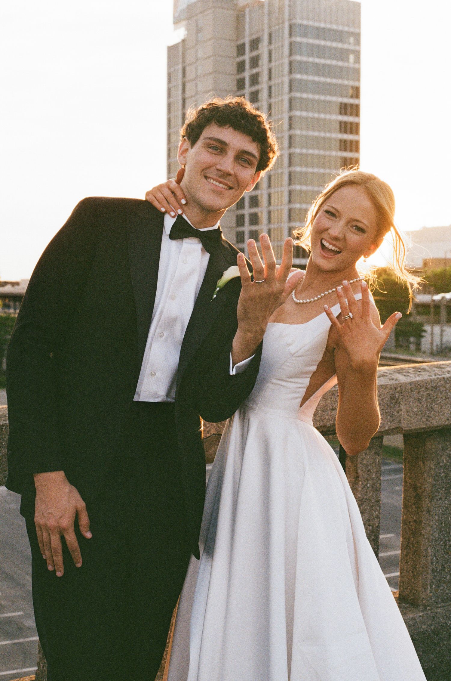 Happy newlyweds show off their wedding rings while posing outdoors at golden hour in the city.