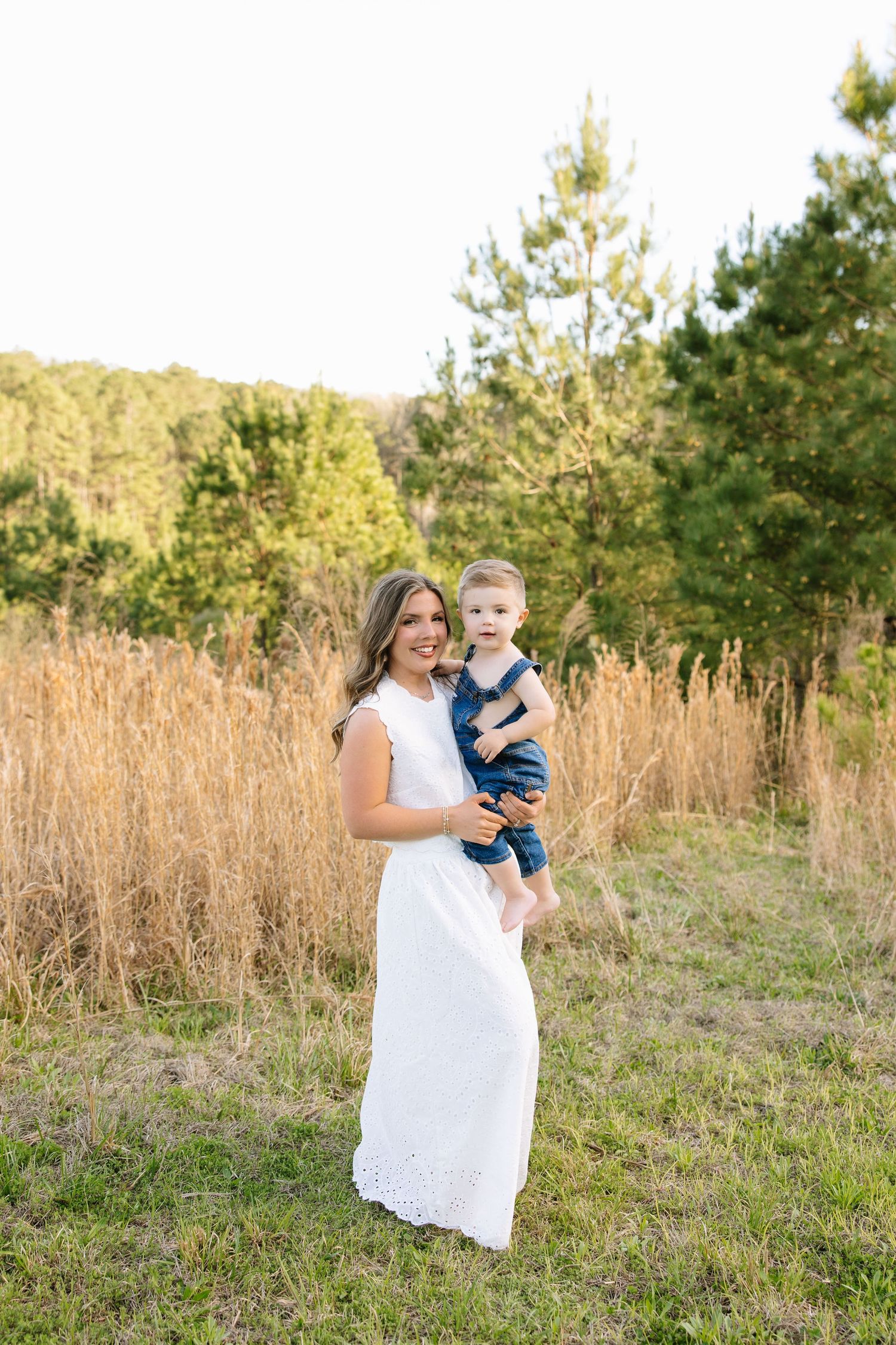 A smiling woman in a white dress holds a happy baby in denim overalls in a sunny field with trees.