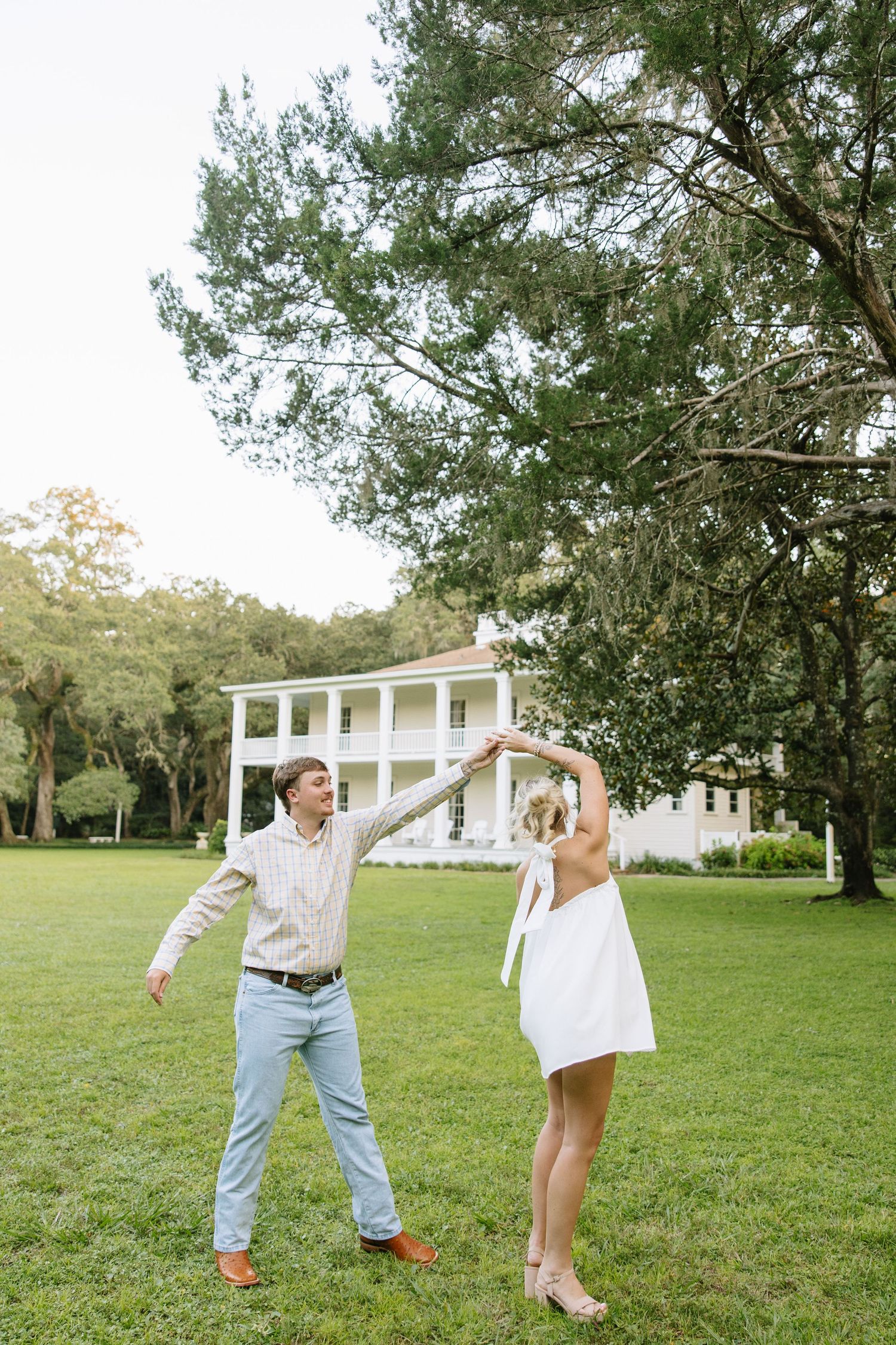 A couple dances on a green lawn in front of a white Southern plantation home surrounded by tall trees.