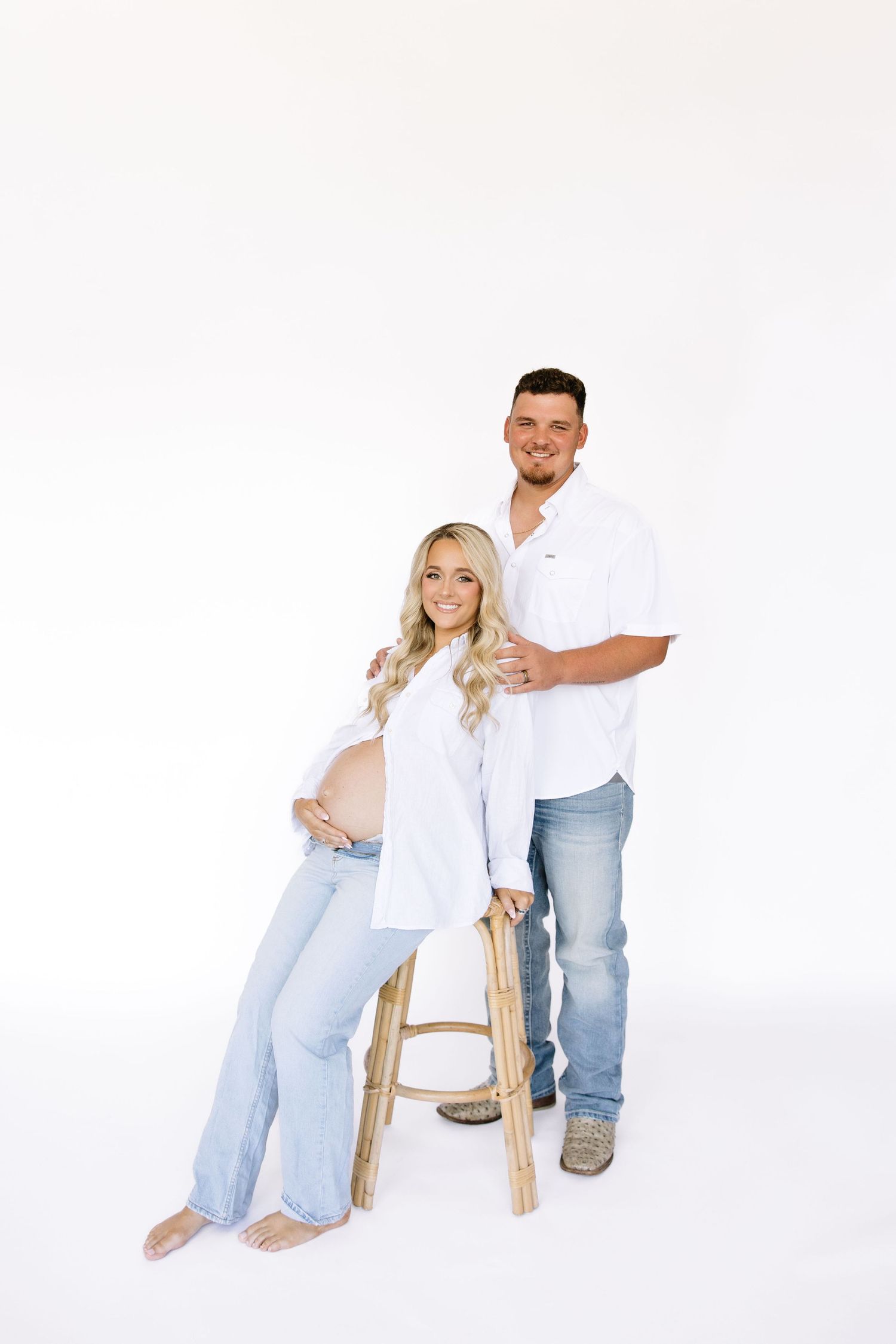Pregnant couple in white shirts and jeans posing in studio.