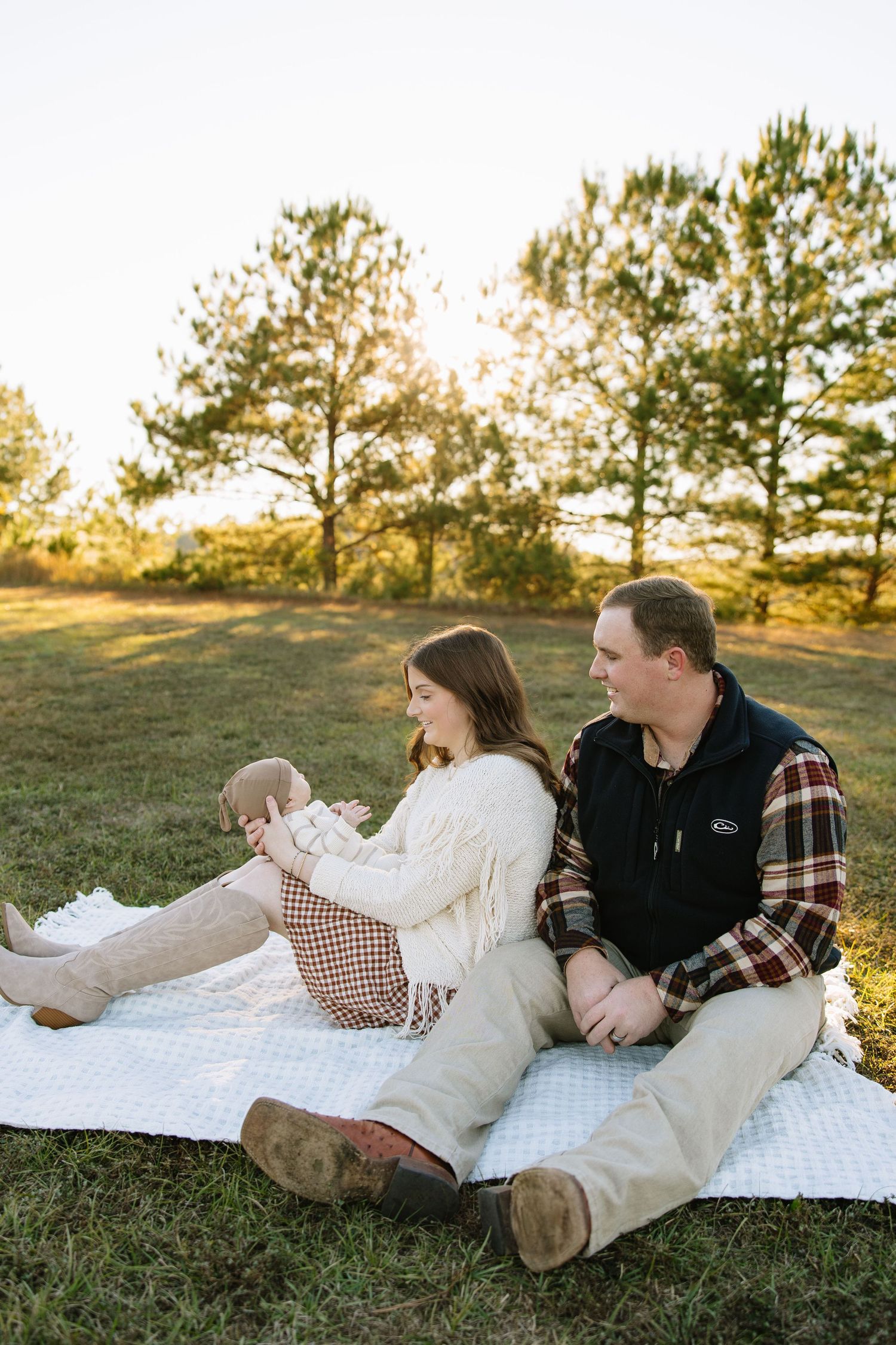 A couple sits on a white blanket in a sunny field, the woman holding a baby while the man looks on warmly.