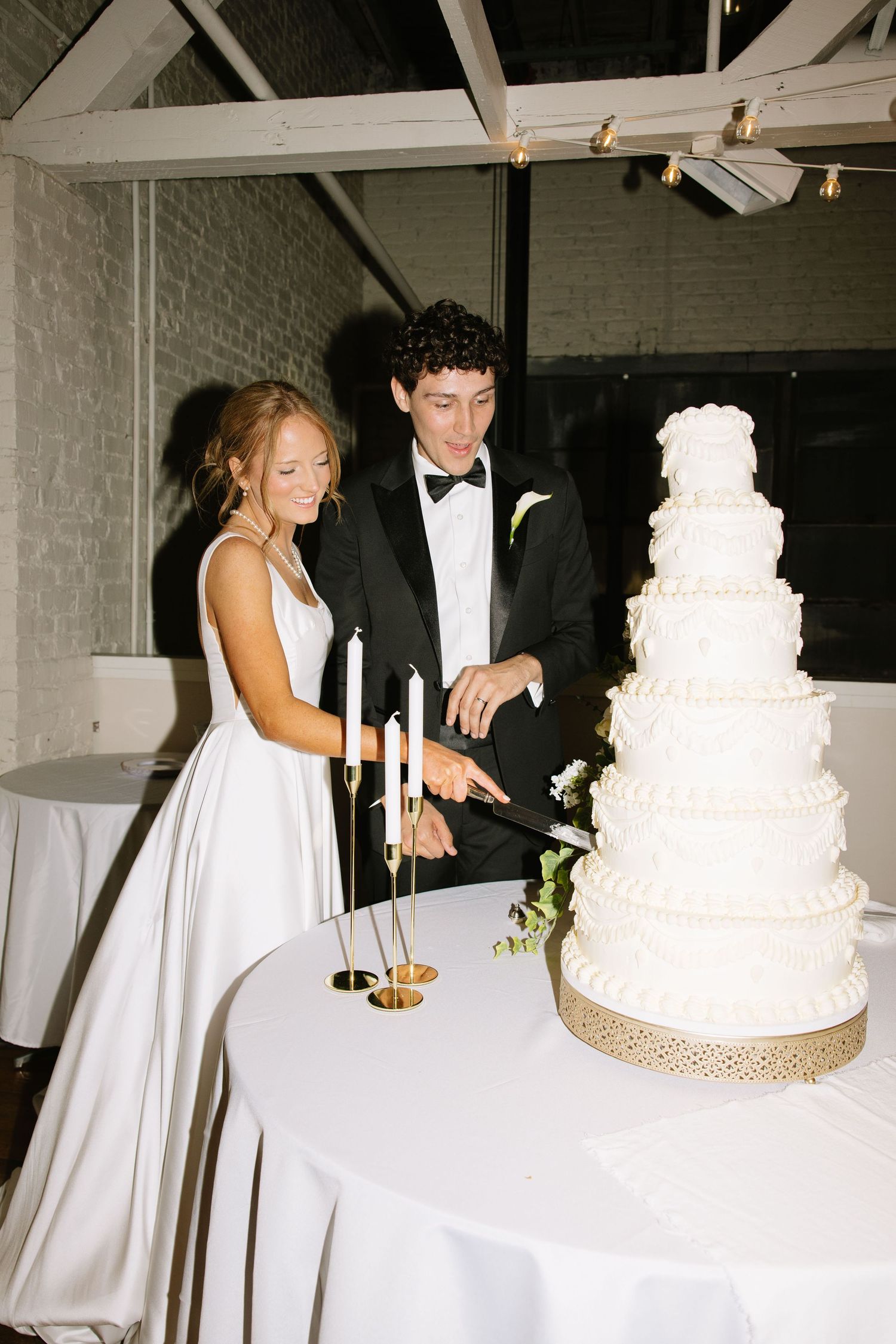 Bride and groom in formal attire cutting a tall five-tier white wedding cake by candlelight indoors.