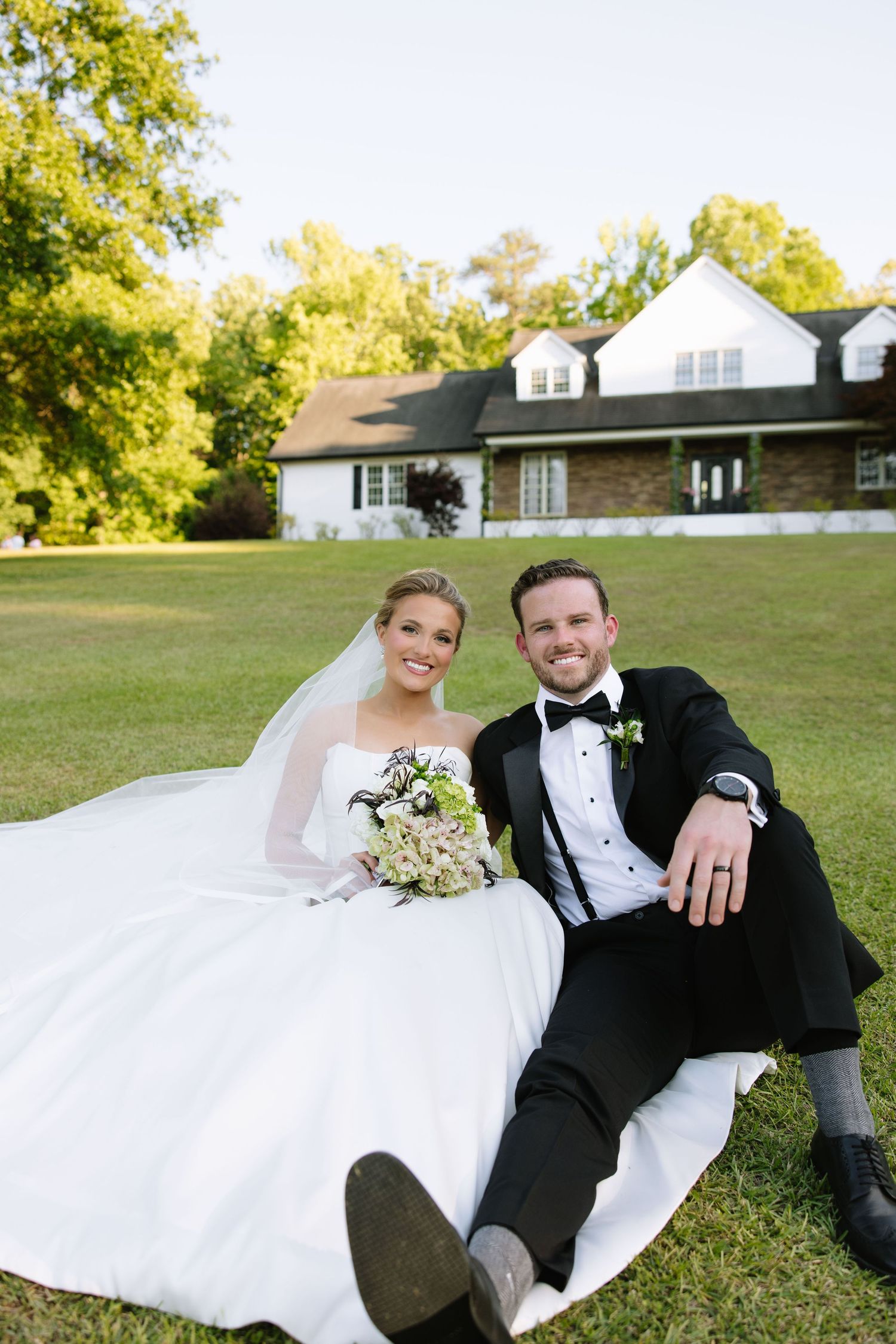 Bride and groom sitting on green lawn in front of white farmhouse, wearing wedding attire with bouquet.