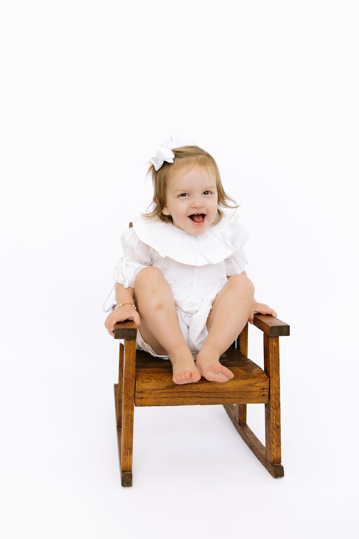 A happy toddler girl in a white romper and bow sits laughing on a small wooden rocking chair against white background.