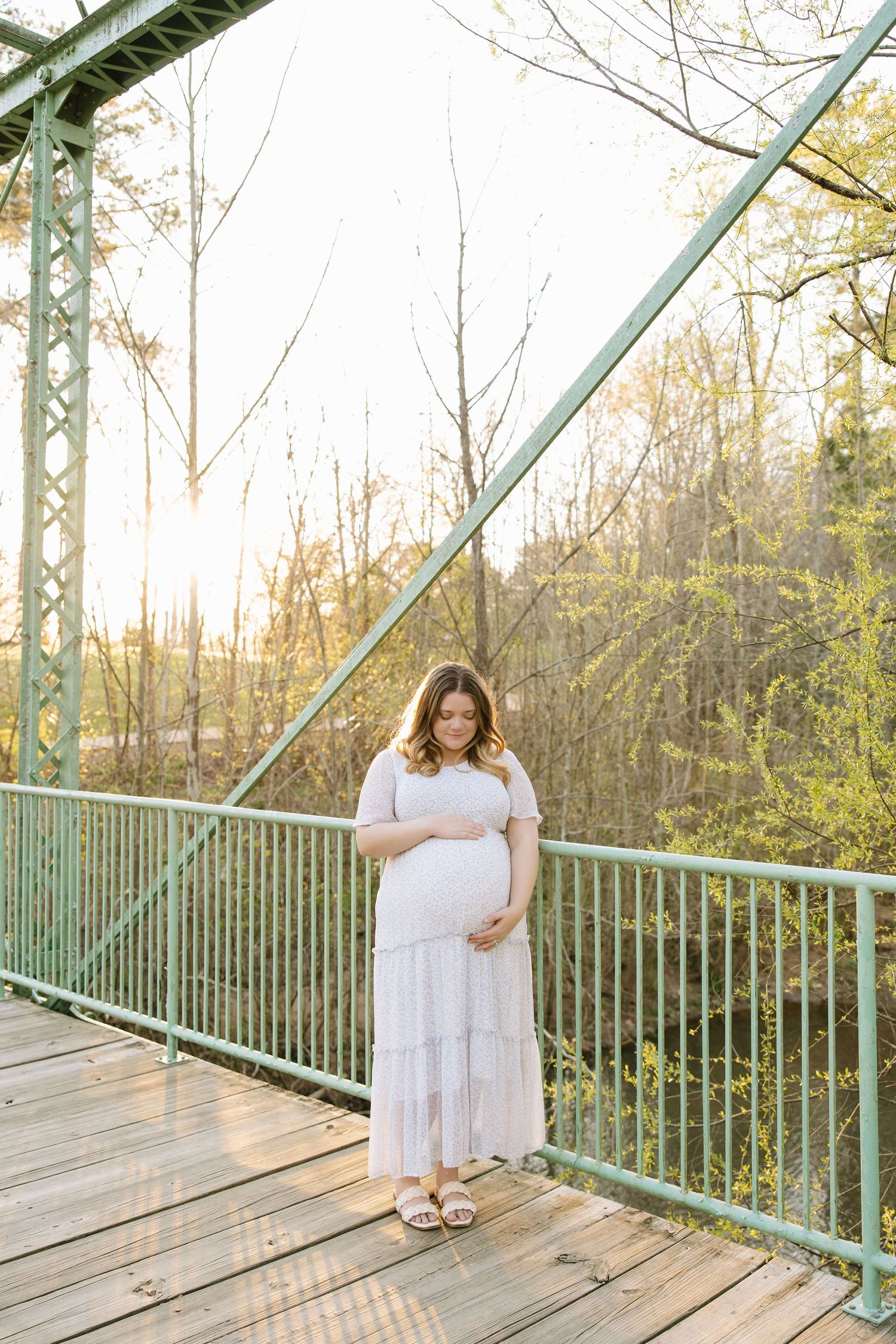 Pregnant woman in white dress cradling baby bump on green bridge surrounded by spring trees at golden hour.