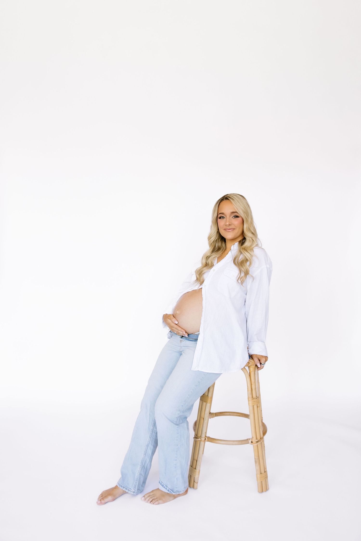 Pregnant woman in white shirt and jeans sitting on wooden stool, cradling baby bump against white background.