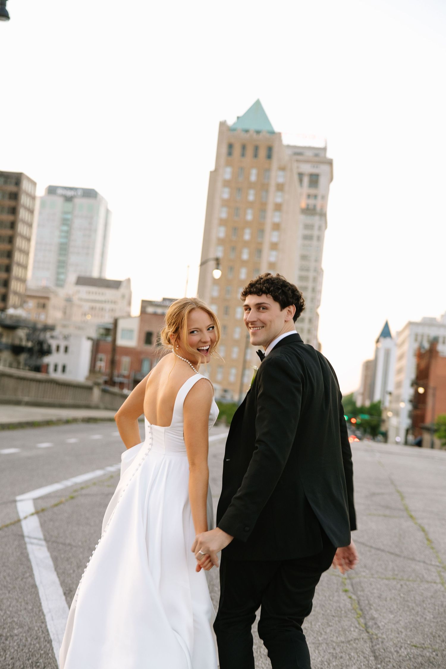 Smiling bride and groom holding hands walking on city street, bride in white gown with open back.