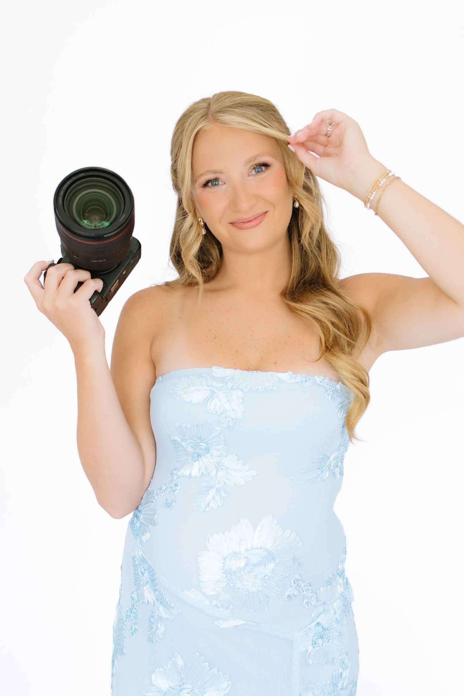 Smiling blonde woman in light blue strapless dress holding a DSLR camera against white background.