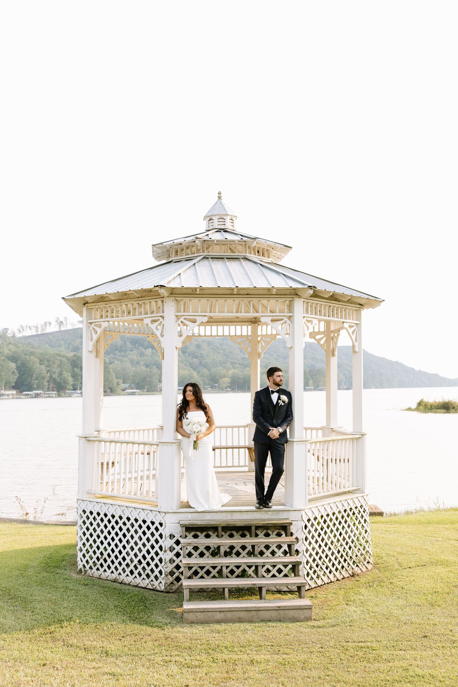White gazebo overlooking a lake serves as a romantic wedding venue with scenic waterfront views.