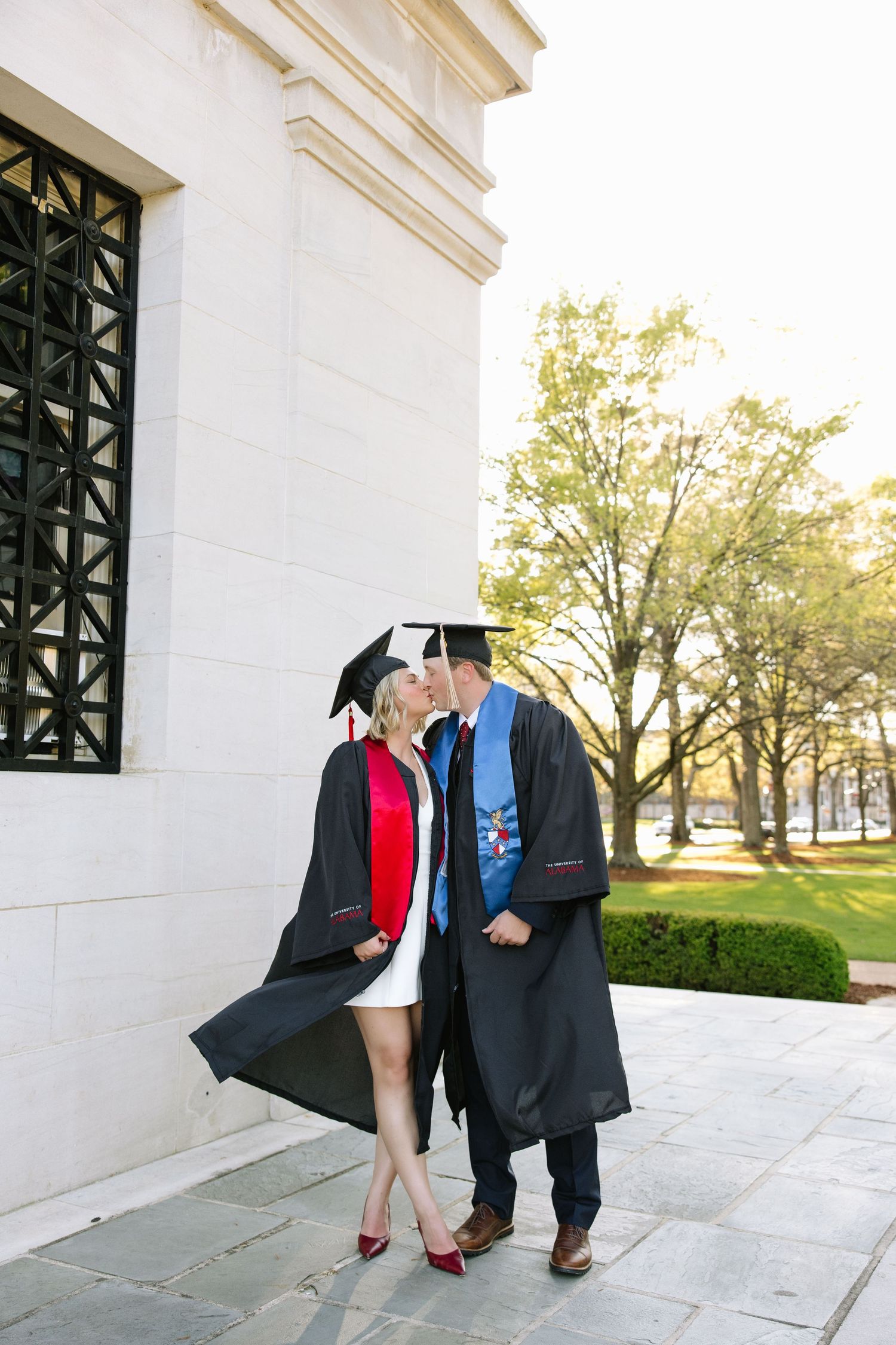 Two graduates in black caps and gowns share a kiss outside a university building on graduation day.