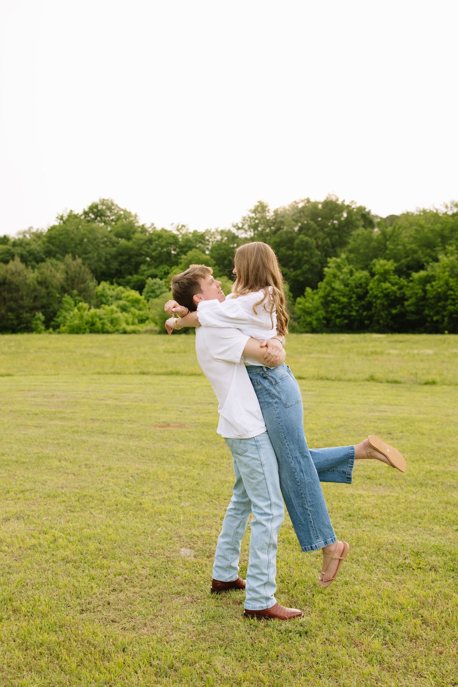A couple embraces and lifts each other joyfully in a lush green field with trees in the background.