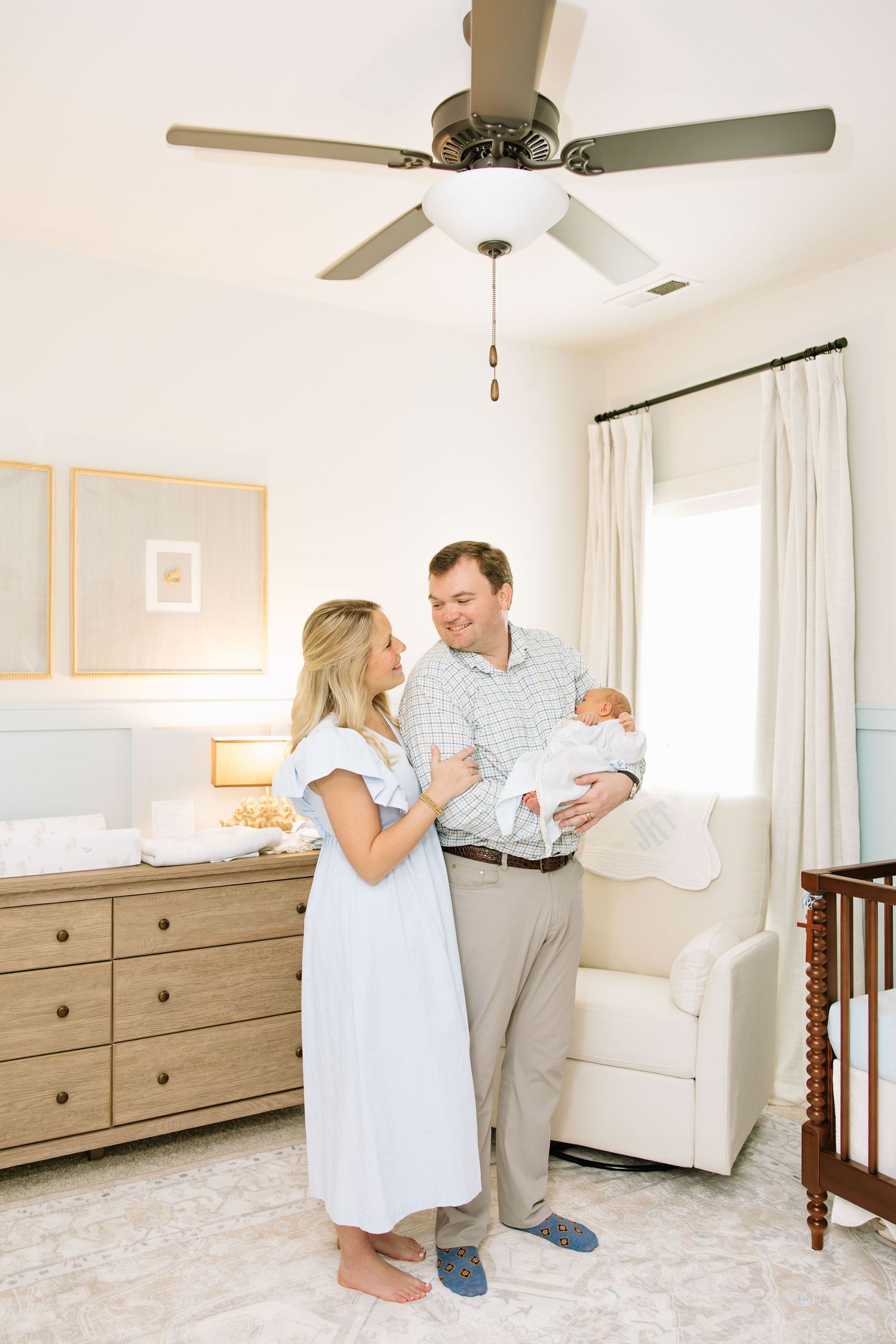 Happy couple standing in a bright nursery, father holding newborn baby, with wooden dresser and crib in background.