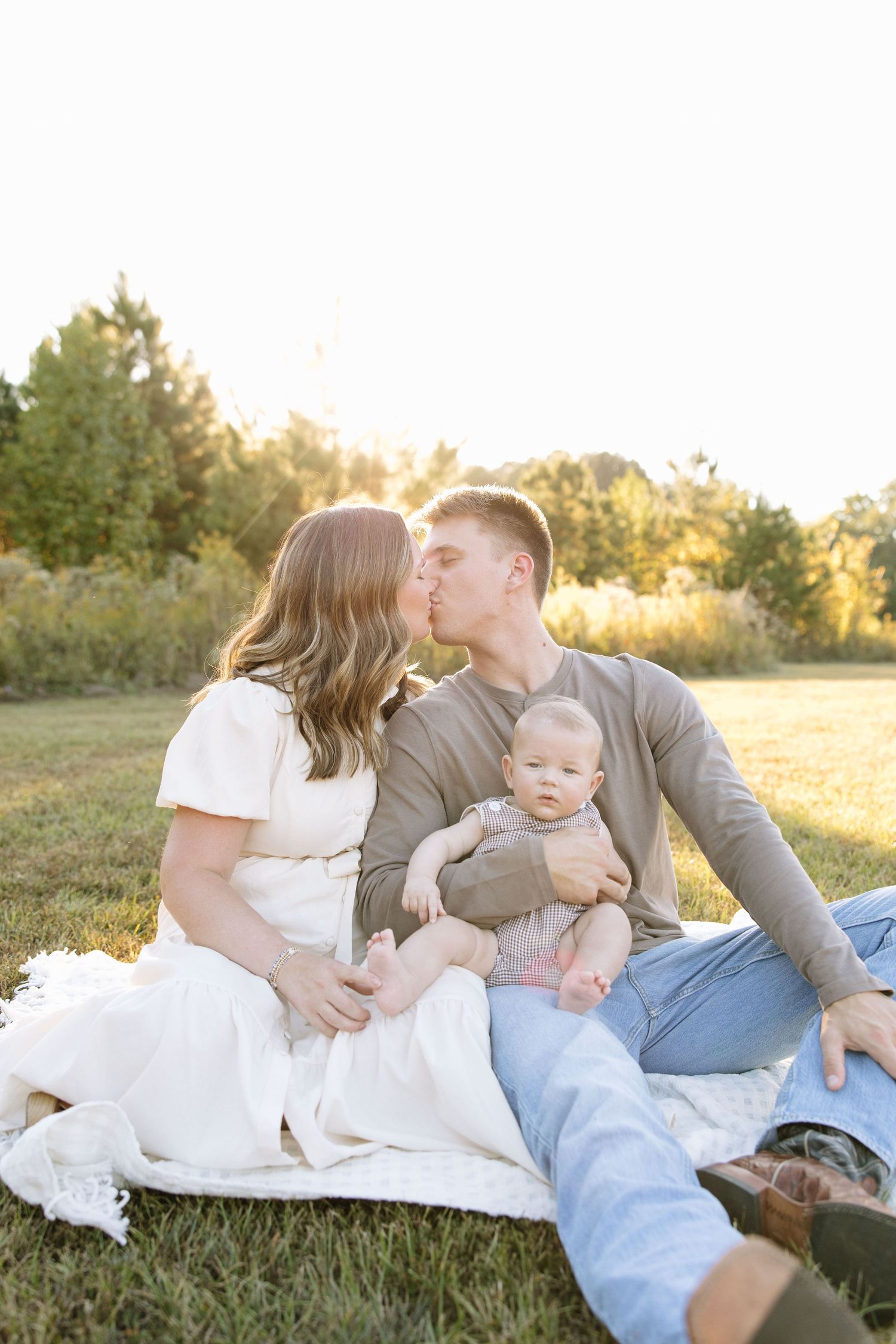 Young couple kissing on a blanket in a sunny field while holding their baby during a golden hour family photo session.