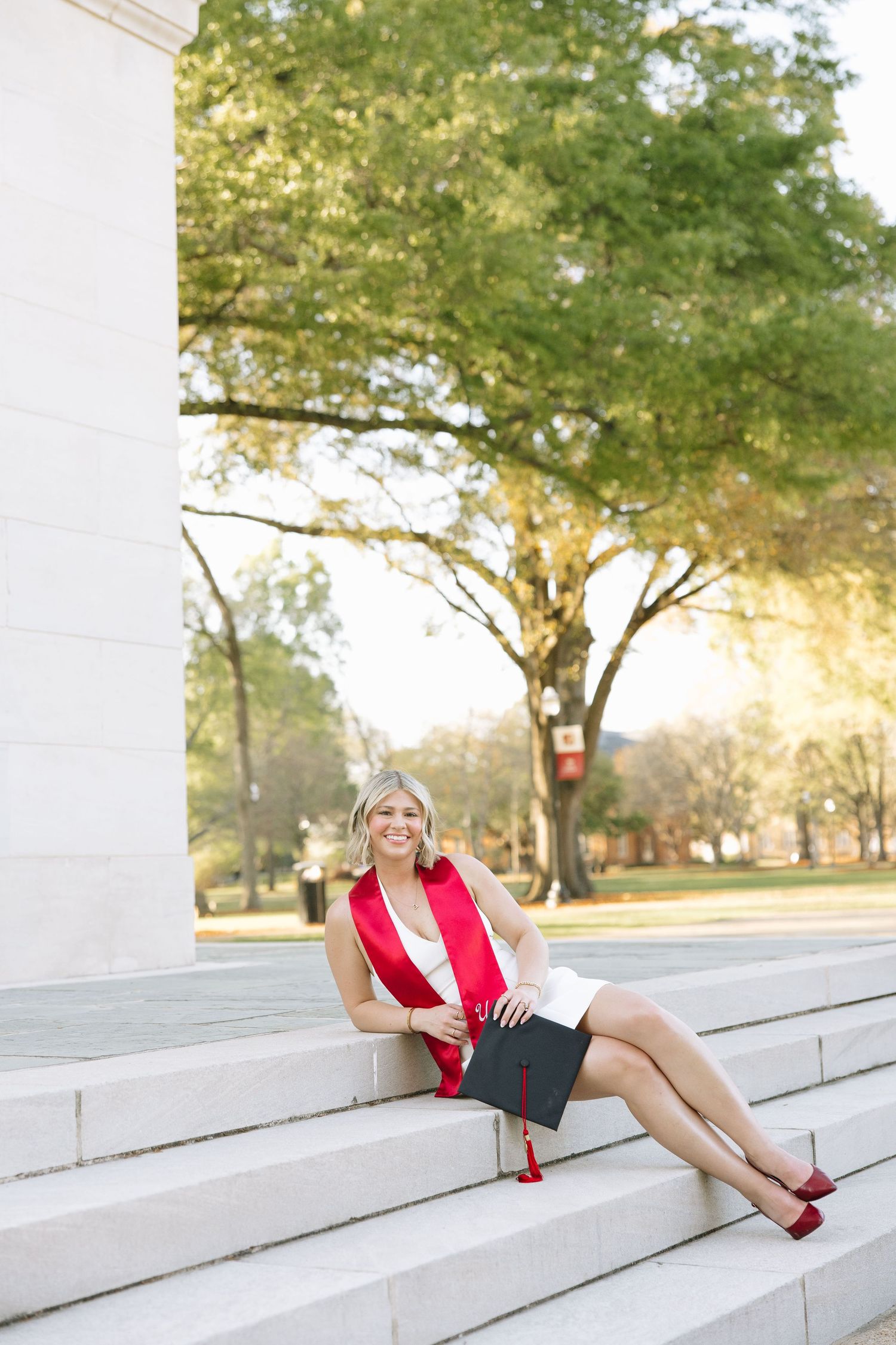 Smiling female graduate in white dress with red sash sits on stone steps holding cap outdoors.