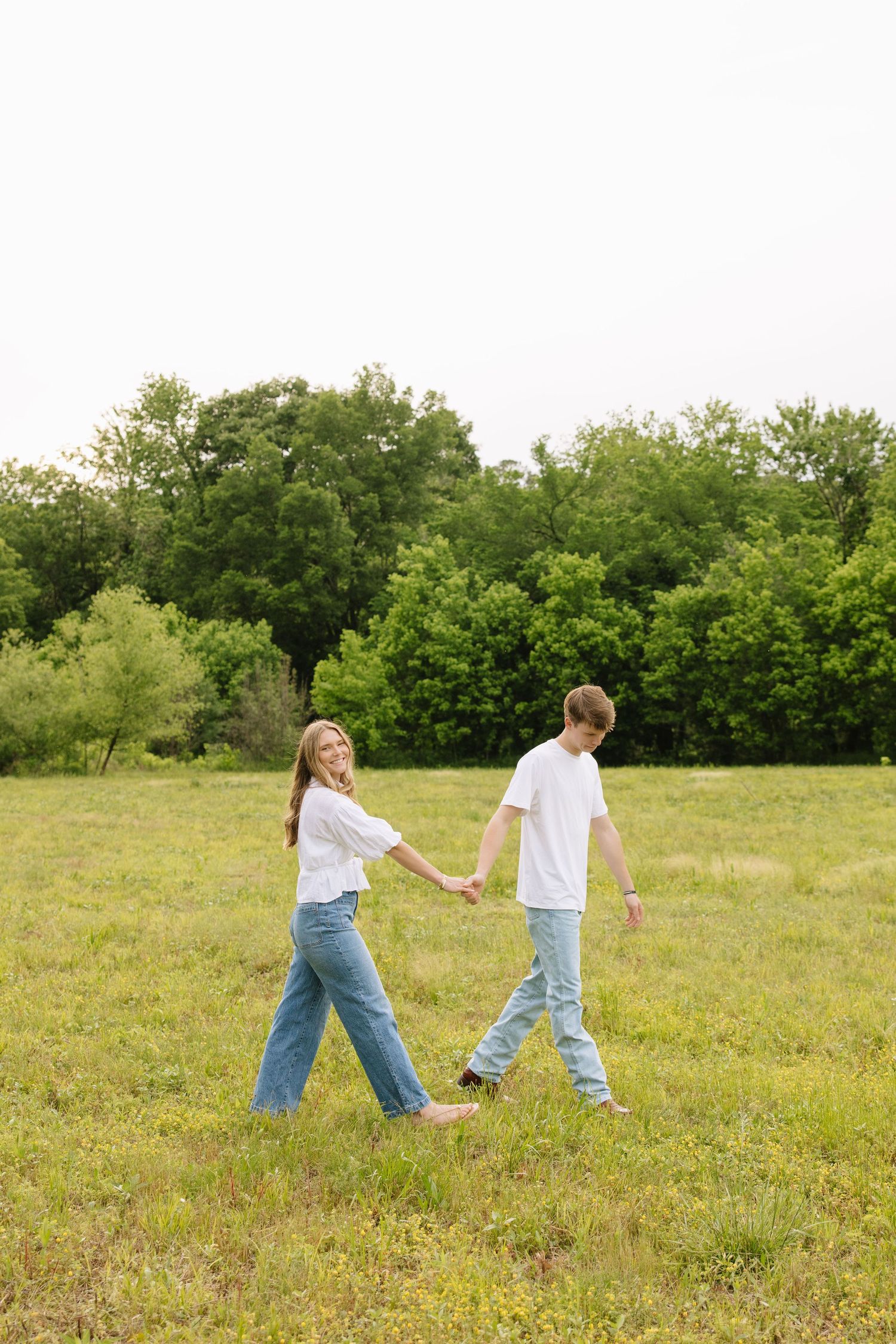 Young couple in white tees and jeans holding hands while walking through a green meadow on a cloudy day.