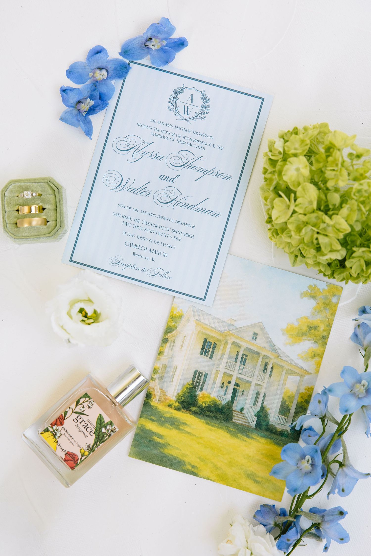 Wedding invitation flatlay with blue florals, green hydrangeas, and a watercolor venue illustration.