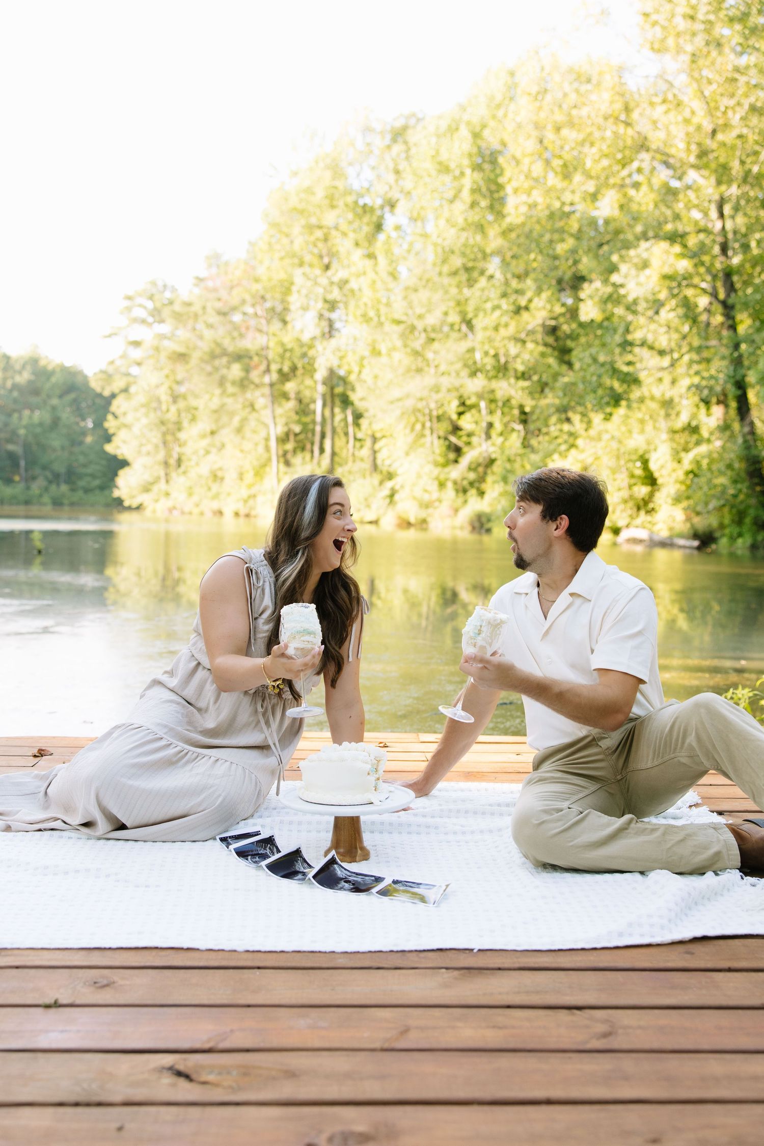 Couple enjoying a romantic lakeside picnic on a wooden dock, sharing drinks and cake surrounded by lush green trees.